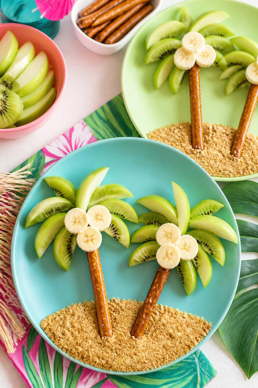Detailed Image Prompt:
A playful summer food craft photo showing pretzel palm trees on plates, pretzel rods used as tree trunks, green apple slices and kiwi pieces arranged as palm leaves, banana slices as coconuts, graham cracker crumb sand at the bottom, bright tropical snack styling, clean kitchen table, no people, no faces, no readable text, no watermark on images.