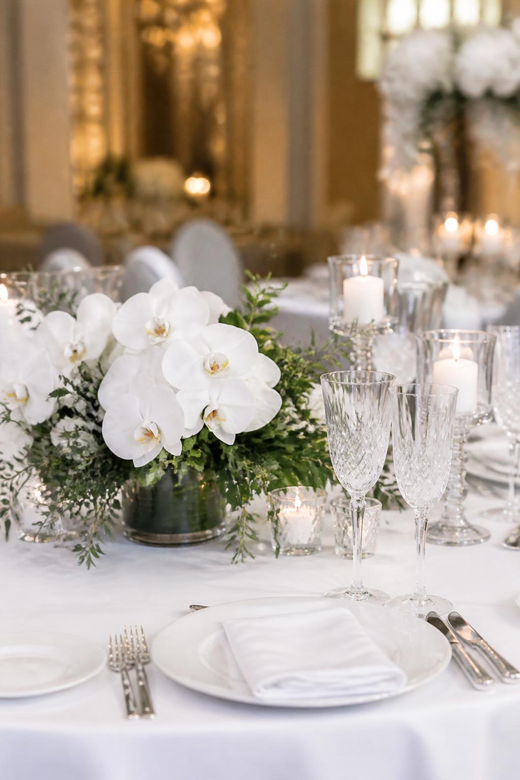 Image Prompt:
A refined wedding table photo showing white orchids in sleek low vases with soft green foliage, paired with white linen, white plates, clear crystal glasses, polished flatware, and elegant candles. Upscale indoor wedding reception setting, no visible faces, no text, no watermark, realistic DSLR editorial photography, (no watermarks on images)