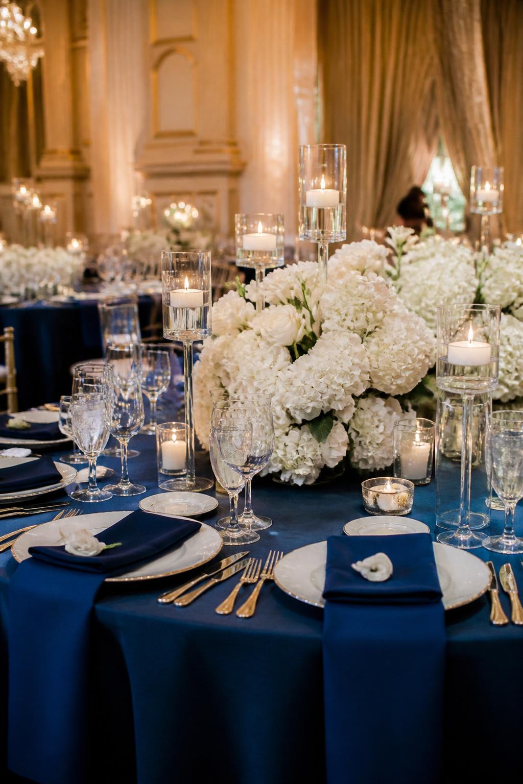 Image Prompt:
A chic wedding table photo showing sapphire blue napkins or runner, white floral centerpieces with roses and hydrangeas, gold or silver flatware, white plates, crystal glasses, and ivory candles in clear glass holders. Formal ballroom or estate reception setting, no visible faces, no text, no watermark, realistic editorial wedding photography, (no watermarks on images)