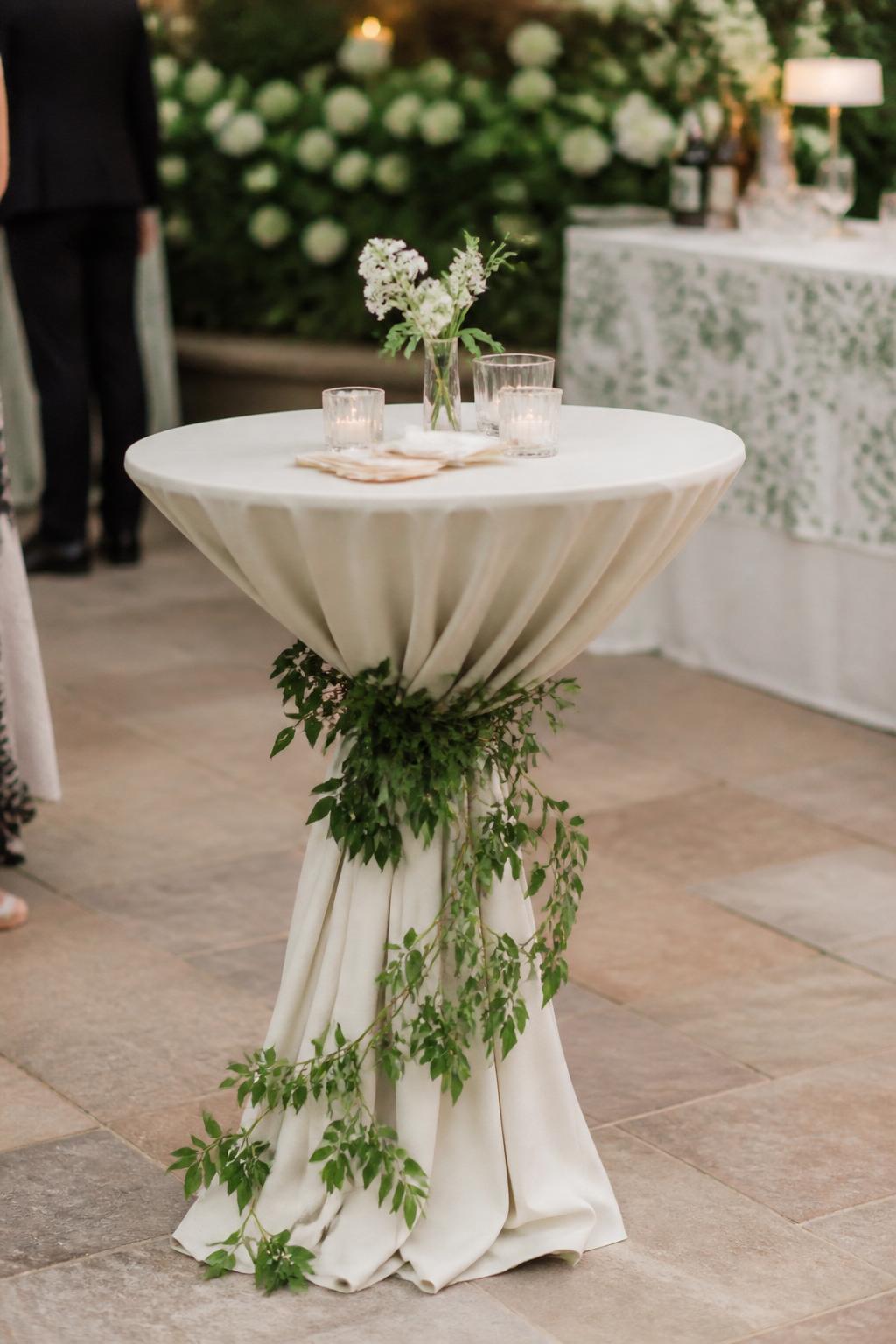 Image Prompt:
A trendy wedding cocktail hour setup showing a high cocktail table with soft greenery wrapped lightly around the table base or pedestal. The tabletop has a small bud vase, cocktail napkins, and a few clear glasses. Garden or tented reception setting, no visible faces, no text, no watermark, realistic editorial wedding photography, (no watermarks on images)