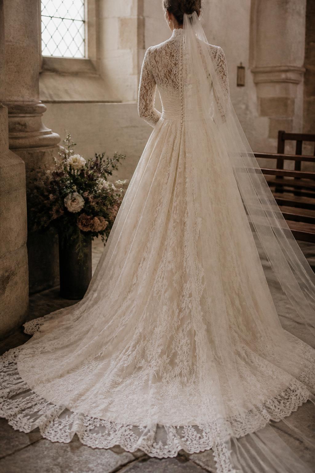 Image Prompt: A luxury bridal editorial image of a bride in a vintage-inspired modest lace wedding dress with long sleeves, high neckline, and delicate lace details, standing in an old stone church or chapel. Show only from the neck down or from behind with the head turned away. Include antique church textures, soft muted florals, timeless styling, realistic lace detail, elegant posture, premium wedding magazine feel, no visible face, no text, no watermark. (no watermarks on images)