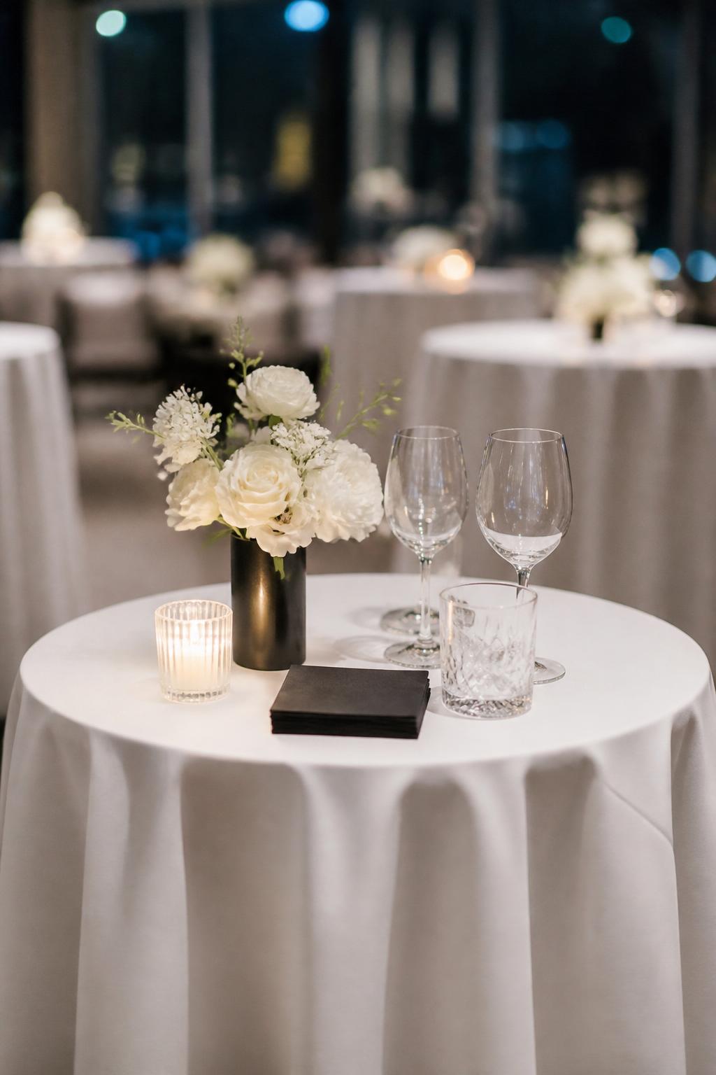 Image Prompt:
A sleek modern wedding cocktail table with a crisp white tablecloth, a small black vase holding white flowers, black cocktail napkins, clear glassware, and soft reception lighting. Clean city wedding venue, no visible faces, no text, no watermark, realistic DSLR wedding photography, (no watermarks on images)