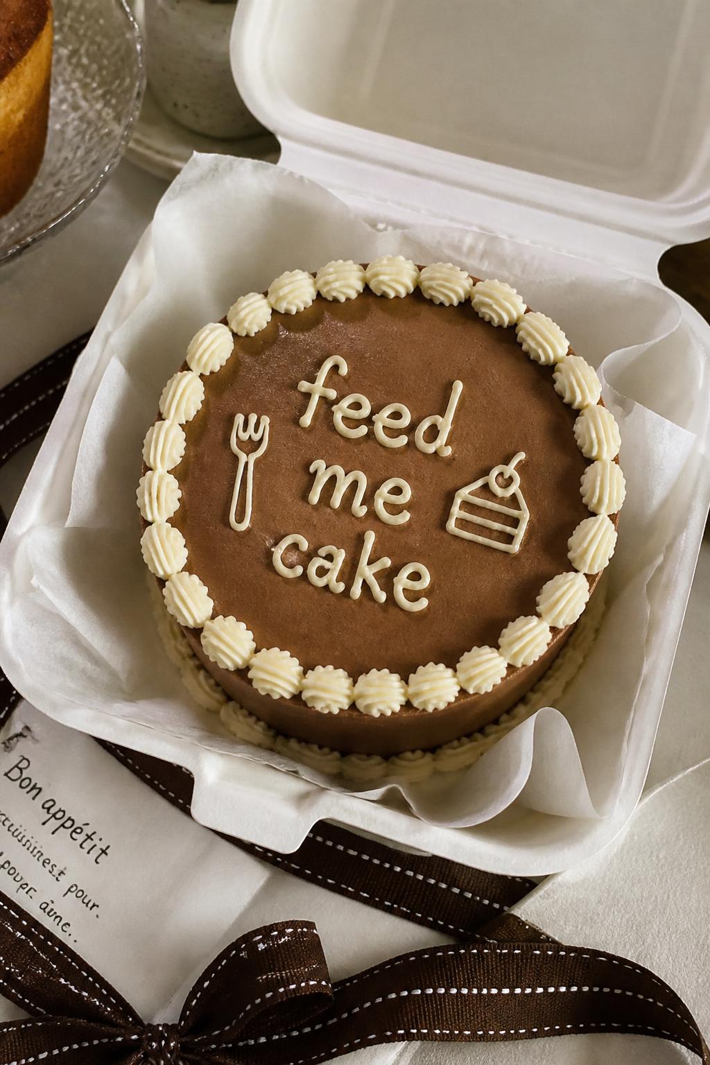 Image Prompt:
A funny bento cake inside an open white cake box, chocolate brown frosting, cream piped border, handwritten icing text reading “feed me cake,” tiny fork and cake slice doodles in icing, cozy dessert table styling, realistic DSLR food photography, no people, no hands, no faces, no watermarks on images