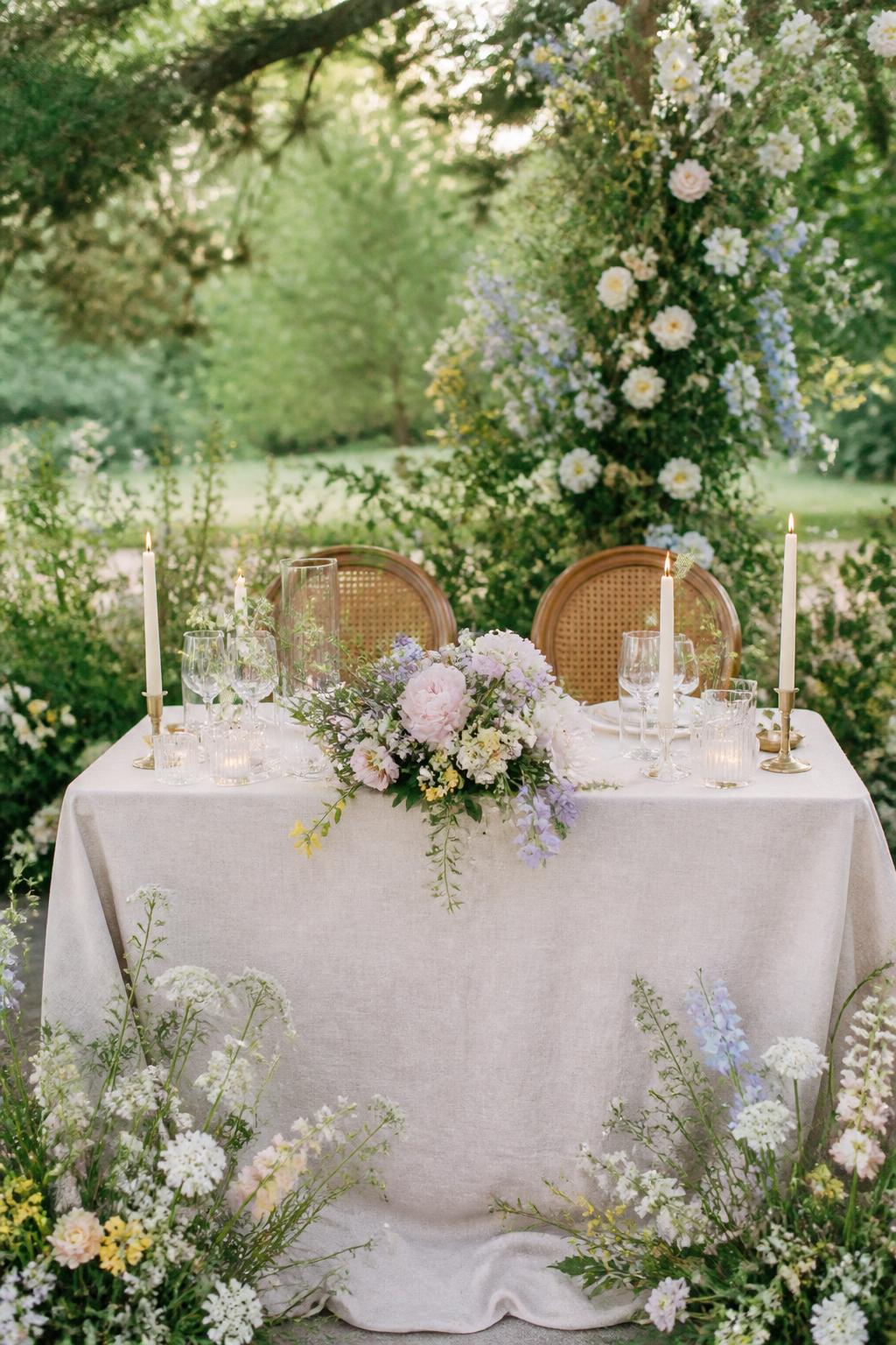 Image Prompt:
A pretty garden wedding sweetheart table styled with loose wildflower arrangements in white, pale yellow, blush, lavender, and soft blue tones, natural linen, taper candles, and clear glassware. Outdoor or garden reception setting, no visible faces, no full people, realistic editorial wedding photography, no text, (no watermarks on images)