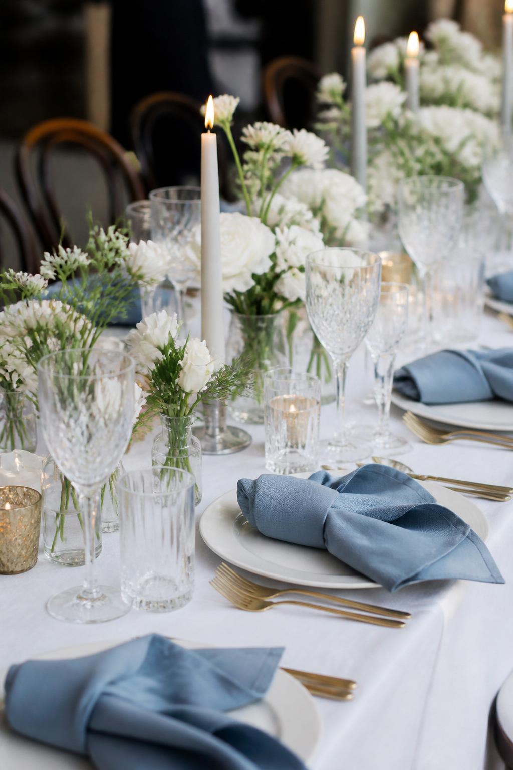 Image Prompt:
A charming civil wedding table styled with dusty blue linen napkins, white plates, small white floral arrangements, clear crystal glasses, silver or gold flatware, and soft candlelight. Intimate city wedding lunch or dinner setting, no visible faces, no text, no watermark, realistic editorial wedding photography, (no watermarks on images)