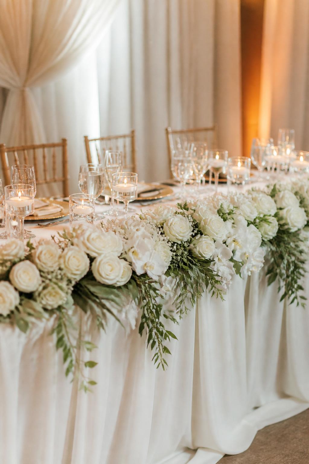 Image Prompt:
A romantic wedding reception photo showing a long white wedding table with soft draped linen and a floral arrangement across the front edge using white roses, white orchids, and delicate greenery. Elegant table settings, candles, clear glassware, and warm refined reception lighting. No visible faces, no full people, no text, no watermark, realistic DSLR wedding photography, (no watermarks on images)