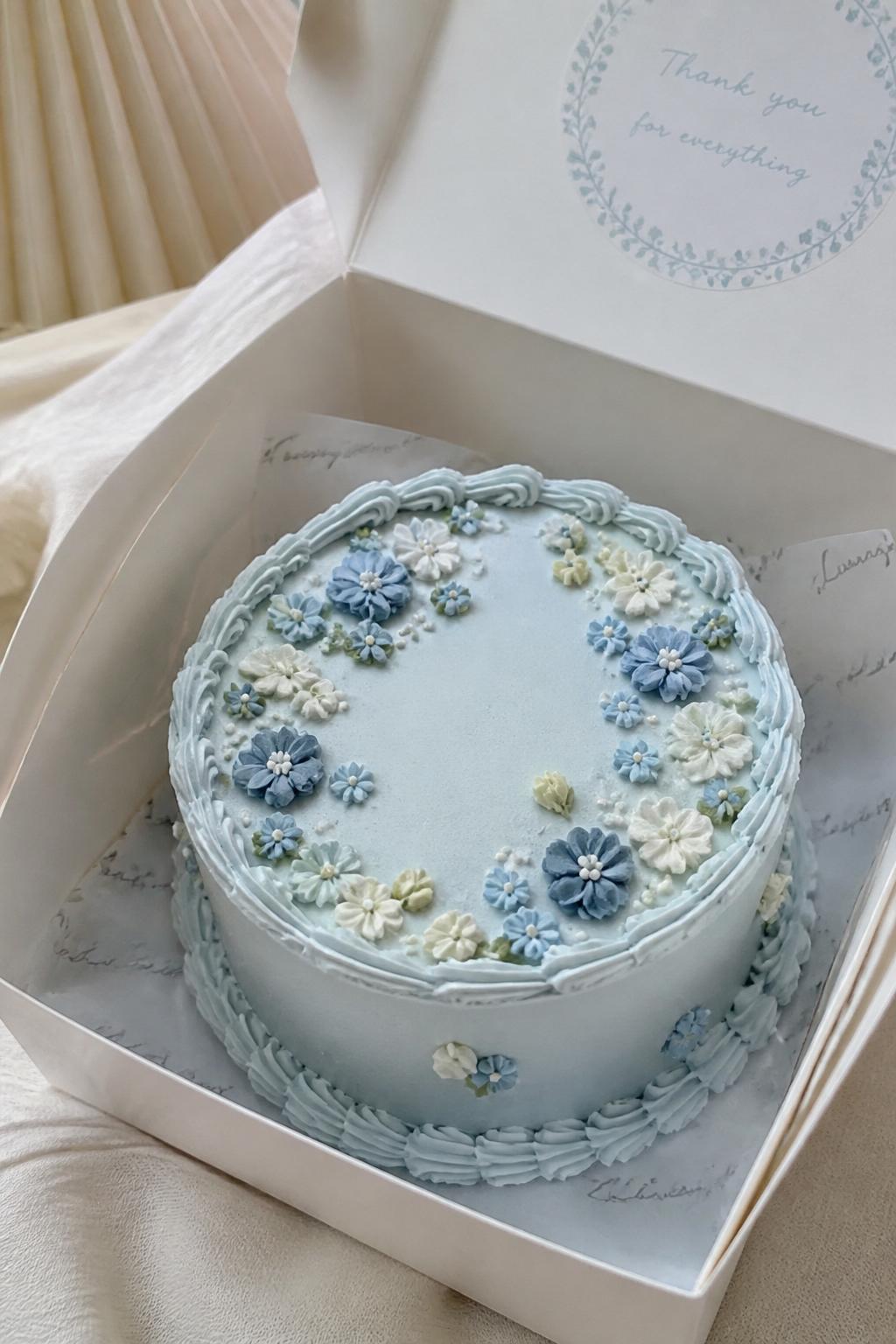 Image Prompt:
A delicate blue floral bento cake inside a white open cake box, smooth pale blue frosting, tiny buttercream flowers in blue, white, and cream tones, elegant piped border, fresh romantic bakery styling, soft natural light, realistic close-up dessert photography, no people, no hands, no faces, (no watermarks on images)