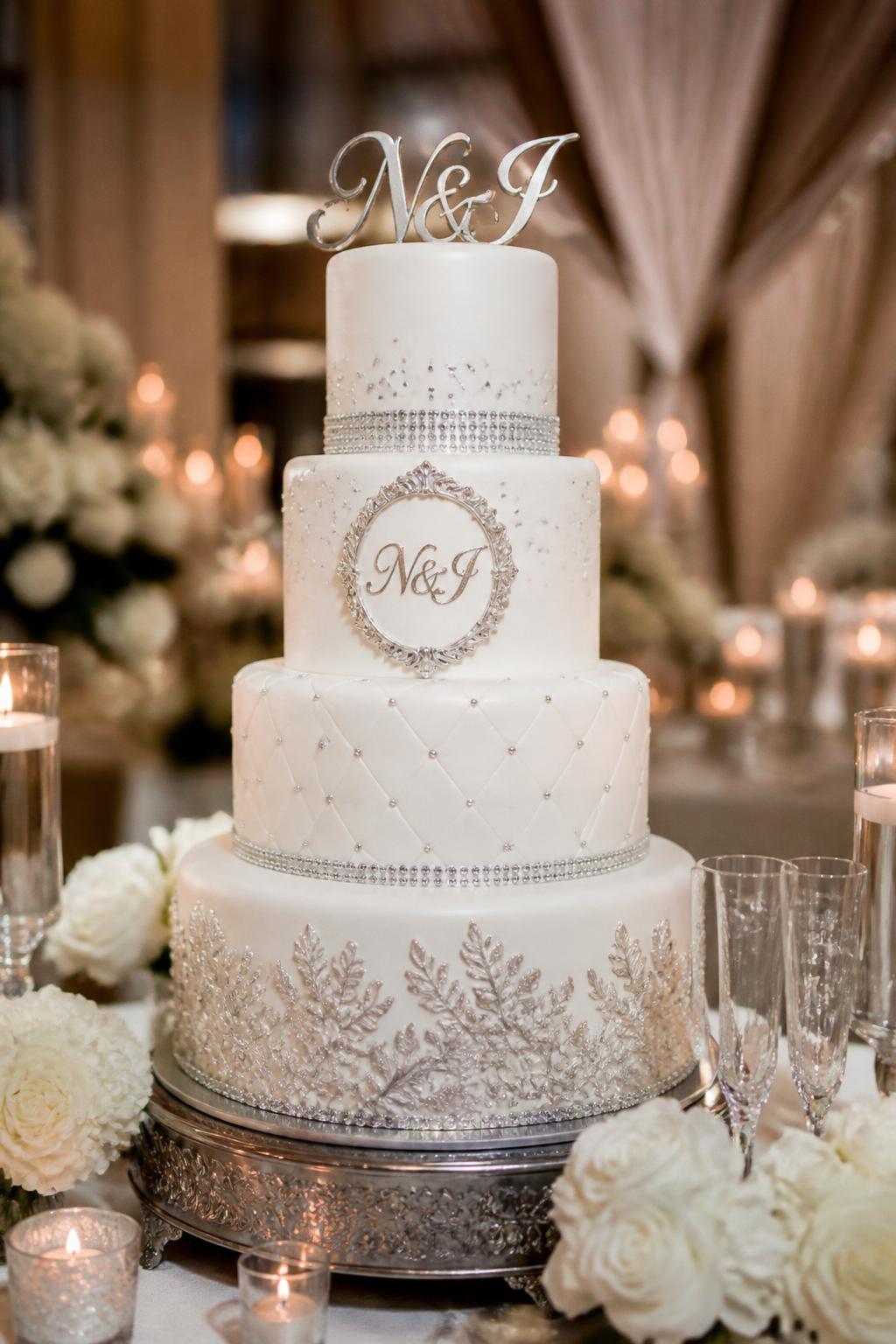 Image Prompt: Ultra-realistic wedding reception photo of a monogram bling wedding cake featuring elegant white tiers, sparkling crystal details, and a stylish monogram plaque or monogram topper as the main design feature. Display the cake at a real wedding party scene with florals, candles, soft table styling, and a luxury reception backdrop. The monogram should look refined and readable but still realistic. No faces visible. If any people appear, show only cropped hands, side silhouettes, or back views. Editorial wedding photography, ultra-detailed, realistic lighting and textures. (no watermarks on images)