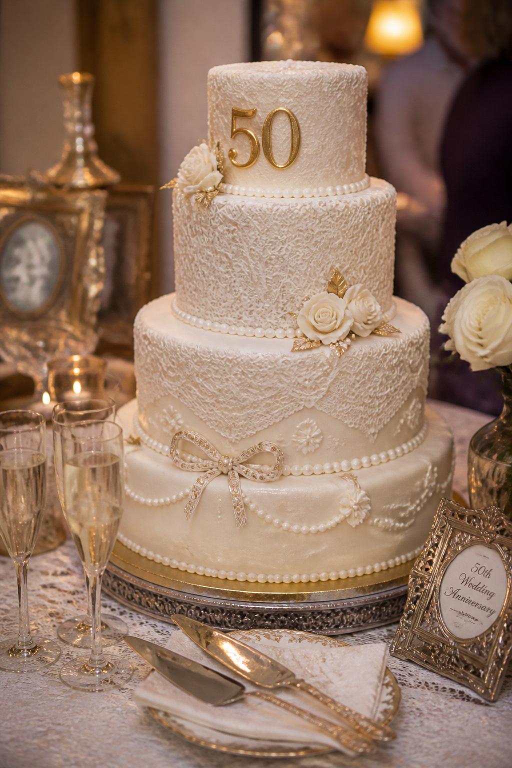 Image Prompt: Ultra-realistic 50th wedding anniversary celebration photo of a vintage lace anniversary cake with ivory tiers, lace-inspired icing detail, delicate piping, pearl accents, and soft gold touches. Place the cake on a decorated dessert table with candles, champagne flutes, antique-style decor, white roses, fine linens, and warm indoor celebration lighting. Keep the cake sharp and detailed with the background softly blurred. No visible faces. If people appear, show only side views, back views, cropped hands, or neck-down figures. Realistic piped lace texture, romantic DSLR event photography, elegant and believable. (no watermarks on images)