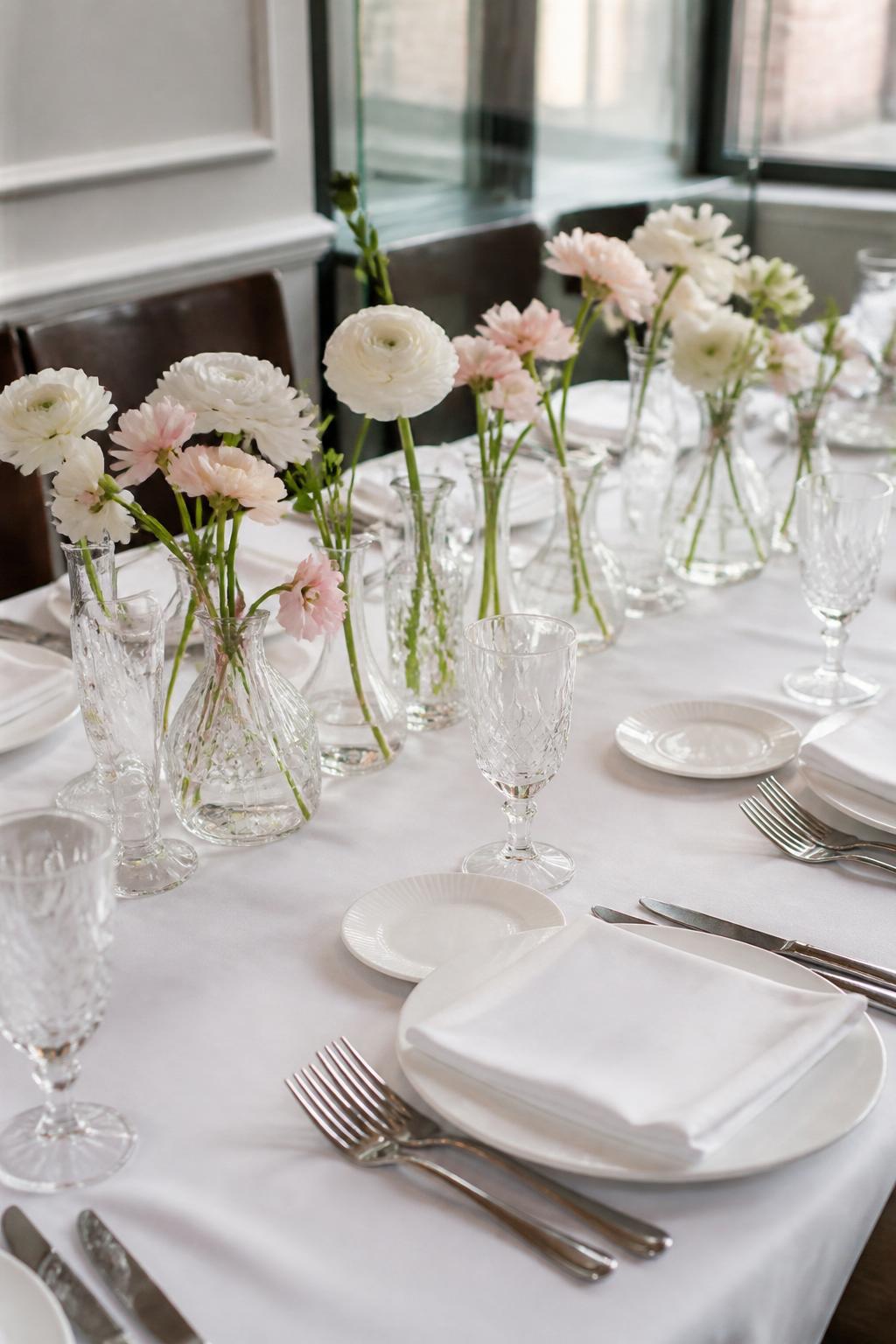 Image Prompt:
A professional editorial civil wedding reception photo showing a small elegant white table styled with a neat row of clear bud vases, each holding one or two delicate flowers in white and blush tones. Add crisp white linen, white plates, clear crystal glasses, folded linen napkins, polished flatware, and soft natural light. Intimate restaurant or courthouse wedding lunch setting, no visible faces, no full people, table-focused composition, realistic DSLR wedding photography, no text, no watermark, (no watermarks on images)