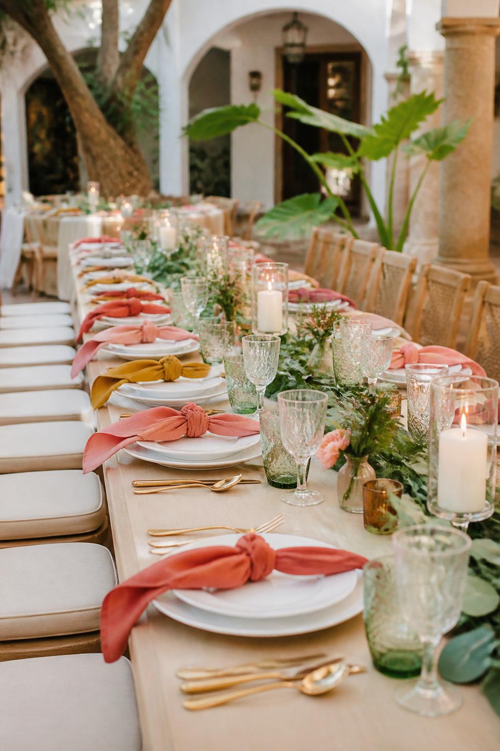 Image Prompt:
A clean modern Mexican wedding table photo showing a long table with simple greenery down the center, colorful linen napkins in coral, mustard, pink, and terracotta tones, white plates, clear glassware, gold flatware, and small candles. The setting is a bright courtyard reception with no visible faces, no text, no watermark, professional editorial photography.