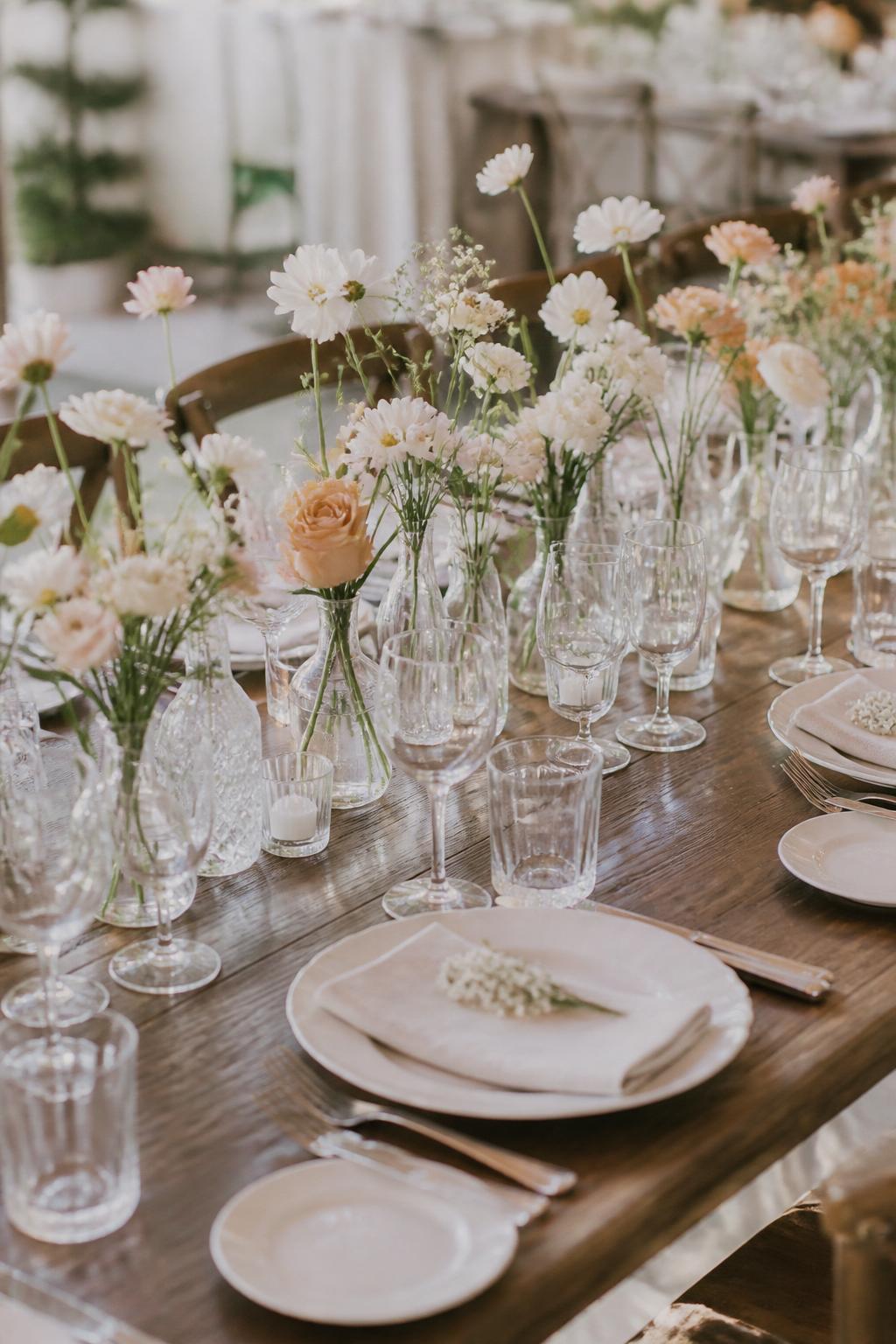 A charming wedding reception photo showing a wooden table lined with small clear bud vases, each holding seasonal stems in white, blush, pale yellow, and soft peach tones. Include white plates, folded linen napkins, clear glassware, and natural elegant styling. No visible faces, no text, no watermark, realistic editorial wedding photography, (no watermarks on images)