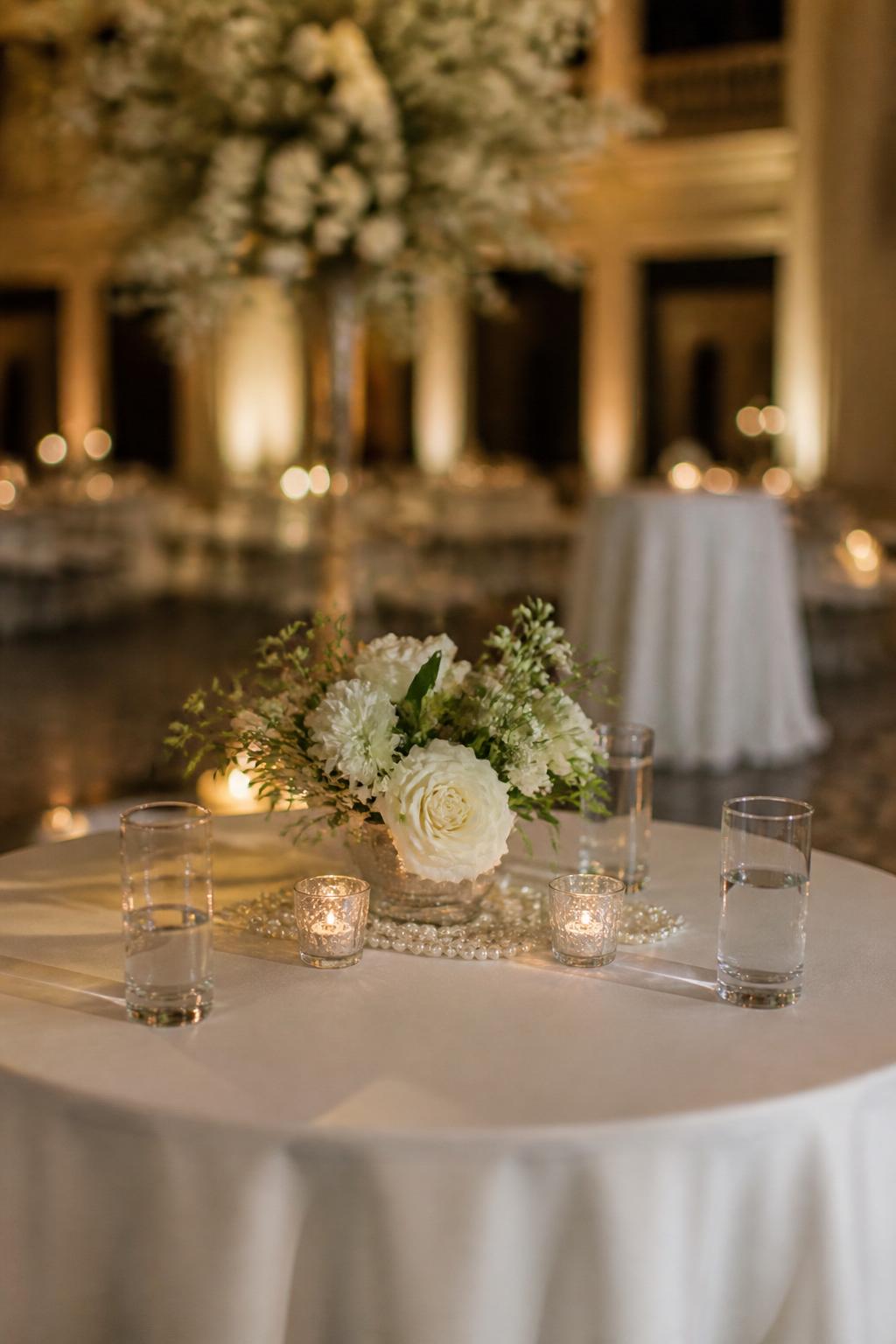 Image Prompt:
A classy editorial wedding cocktail table image showing a small round table with white linen, a tiny white floral arrangement, pearl-like decorative accents placed neatly around the vase, clear glasses, and soft candlelight. Elegant reception setting, no visible faces, no text, no watermark, realistic DSLR wedding photography, (no watermarks on images)