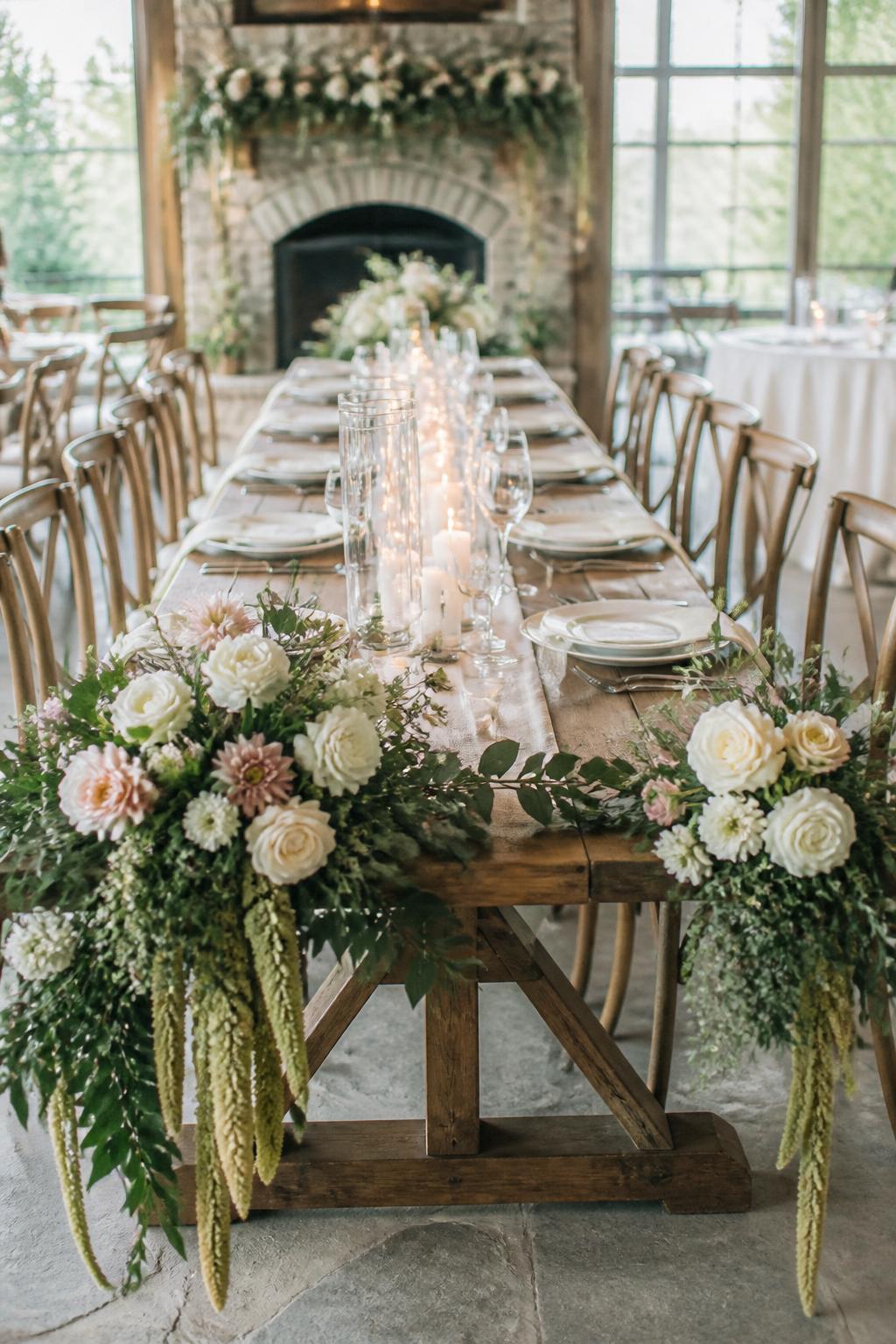 Image Prompt:
A high-end editorial wedding reception photo showing a long wooden farm table with large floral arrangements placed at each end, using white roses, blush flowers, greenery, and trailing stems. The center of the table has candles, elegant place settings, linen napkins, and clear glassware. Rustic elegant wedding venue, no visible faces, no full people, no text, no watermark, realistic DSLR photography, (no watermarks on images)