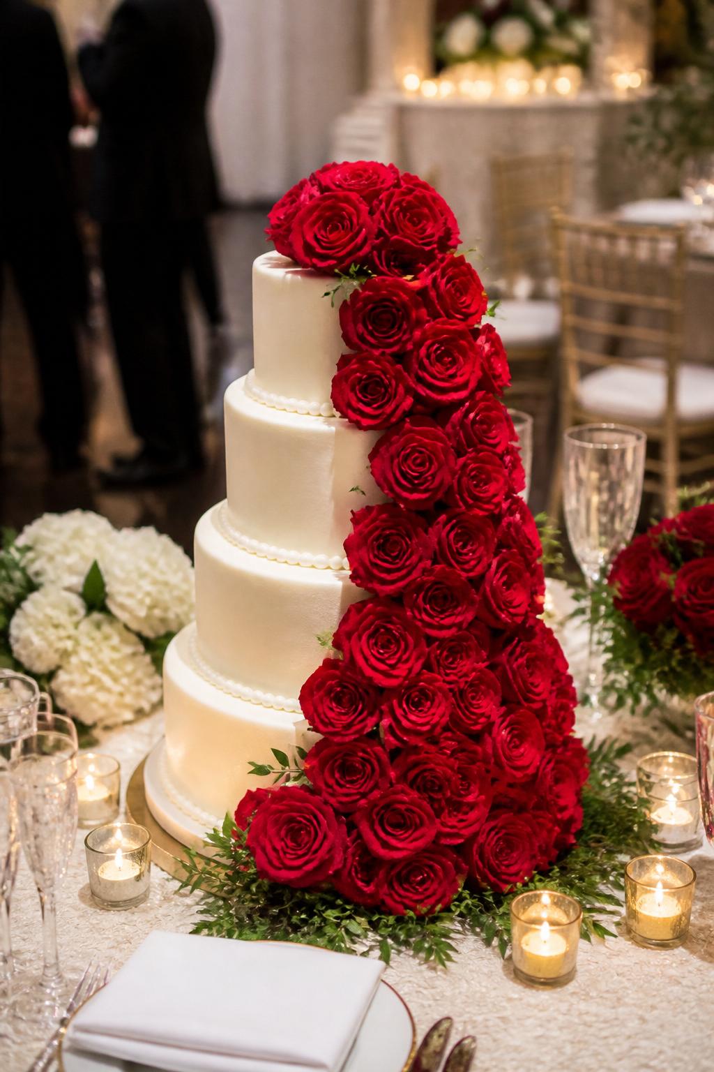 Image Prompt: Ultra-realistic wedding party scene showing a wedding cake with a dramatic cascade of fresh red roses flowing from the top tier to the base. Use smooth ivory or white icing, a few touches of greenery, and a refined romantic finish. Place the cake on an elegant reception cake table with candles, floral centerpieces, fine table settings, champagne glasses, and a softly blurred wedding venue background. Keep the cake in sharp focus. No visible faces. If guests appear, show only side views in blur, back views, cropped torsos, or neck-down figures. Realistic red rose texture, detailed icing, warm romantic lighting, DSLR wedding realism. (no watermarks on images)