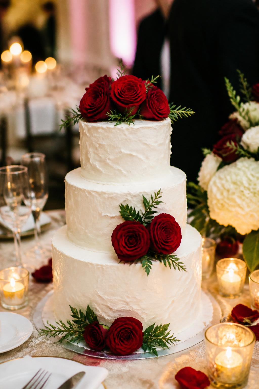 Image Prompt: Ultra-realistic wedding party photo of a textured buttercream wedding cake with soft white or ivory frosting, gentle palette-knife buttercream texture, fresh red roses, and a few delicate greenery accents. Style the cake on a reception cake table with candles, floral centerpieces, glassware, fine linens, and a softly blurred wedding venue backdrop. Keep the cake as the main focus. No visible faces. If guests appear, show only back views, side views in blur, cropped hands, or neck-down figures. Realistic buttercream texture, red rose petals, warm DSLR wedding photography. (no watermarks on images)