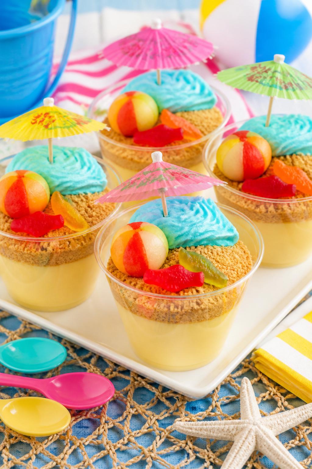 Detailed Image Prompt:
A cute summer food craft photo showing beach pudding cups in clear plastic cups, vanilla pudding topped with crushed graham cracker “sand,” blue-tinted whipped topping as water, tiny fruit beach balls, paper umbrellas, and gummy fish, styled on a bright party table with spoons nearby, playful beach snack look, no people, no faces, no readable text, no watermark on images.