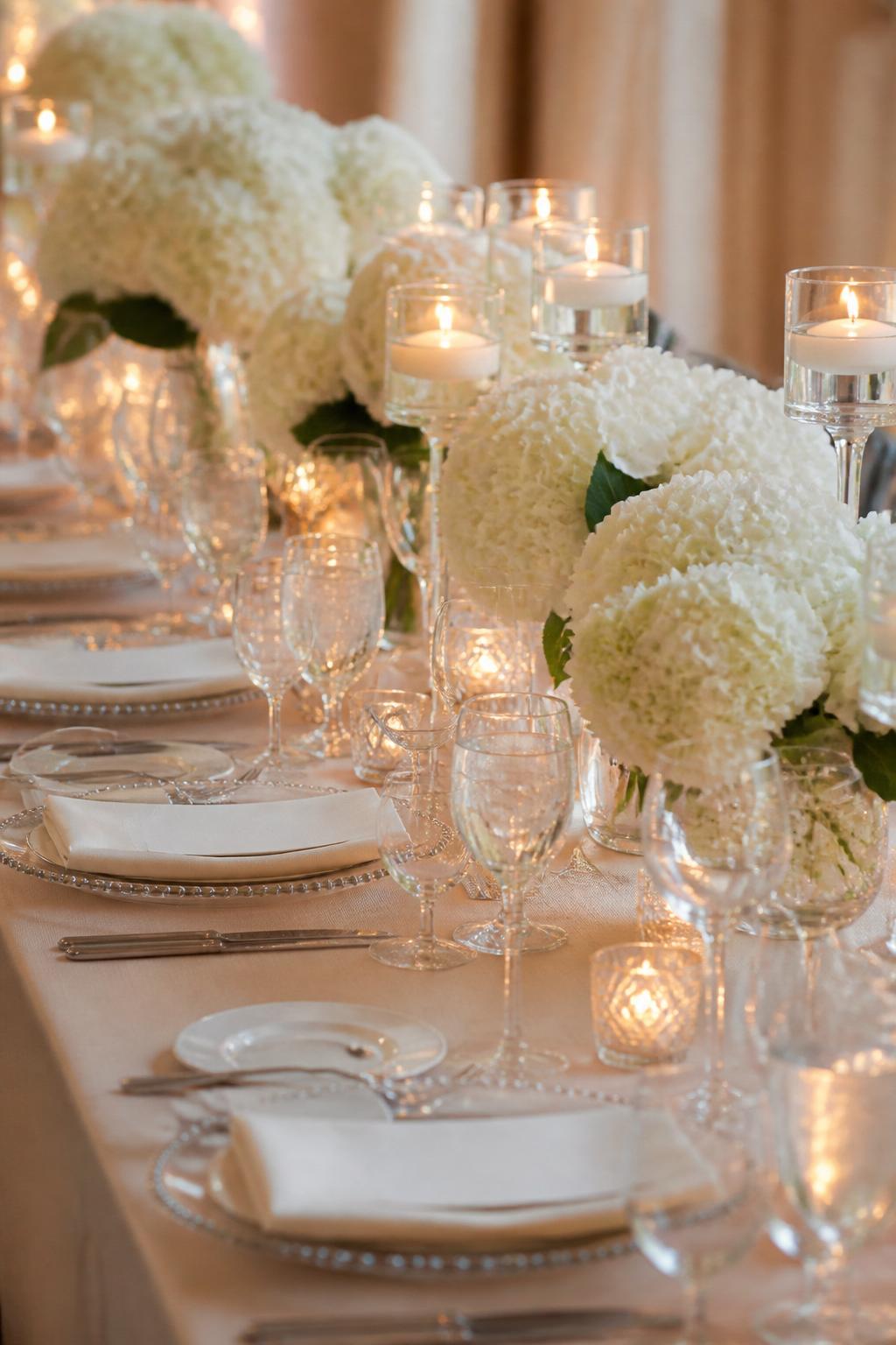 Image Prompt:
A romantic editorial wedding table photo showing white hydrangea centerpieces arranged along a long table, mixed with candle clusters in clear glass votives and elegant holders. The table includes ivory linen, white plates, clear glassware, and soft warm lighting. No visible faces, no text, no watermark, realistic wedding photography, (no watermarks on images)