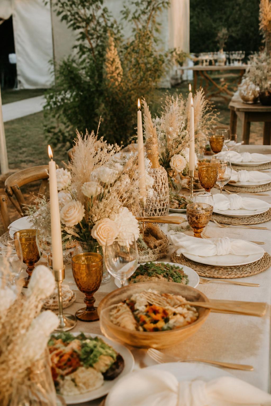 Image Prompt:
A cozy boho family style wedding dinner table with neutral linen, rattan chargers, beige and white flowers, pampas grass used lightly, amber glassware, candles, shared serving dishes, and white plates. Outdoor tented or backyard wedding reception setting, no visible faces, no text, no watermark, realistic DSLR editorial photography, (no watermarks on images)