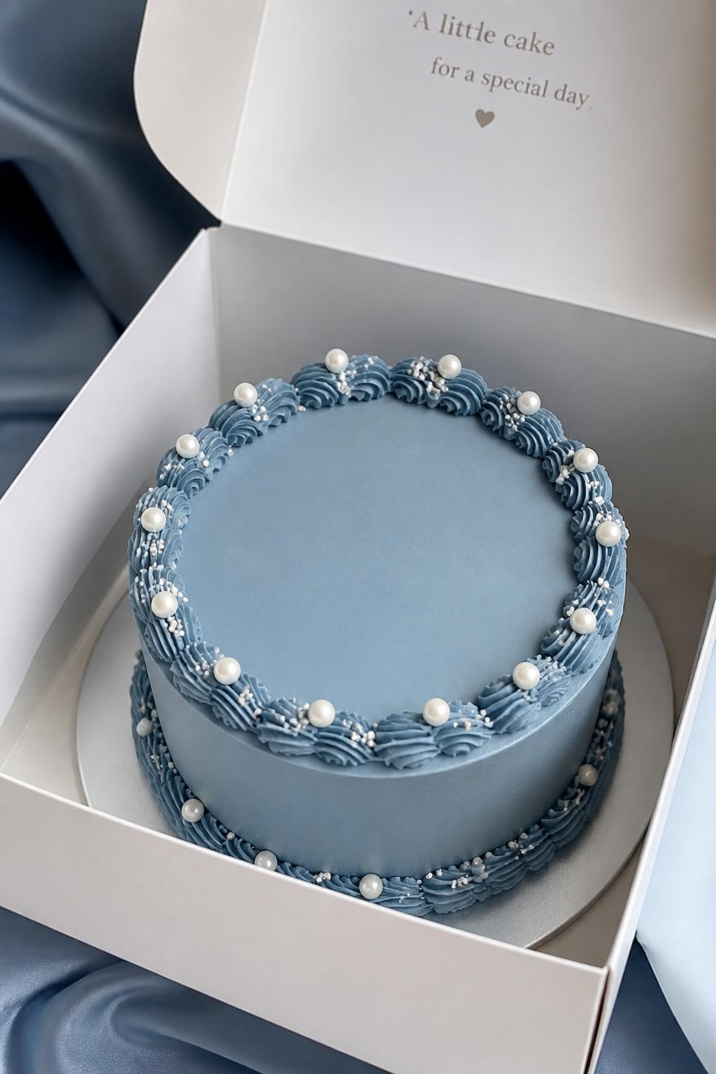 Image Prompt:
A beautiful blue pearl bento cake inside a white cake box, smooth dusty blue frosting, tiny edible pearl accents placed neatly around the top edge, elegant piped border, soft luxury bakery styling, clean backdrop, realistic editorial dessert photography, no people, no hands, no faces, (no watermarks on images)
