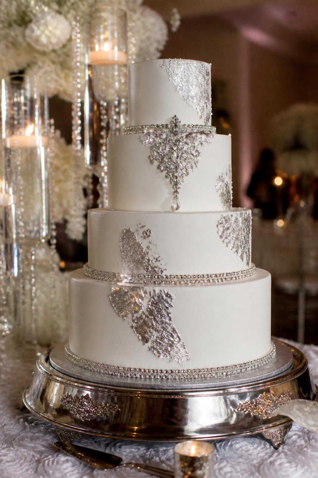 Image Prompt: Ultra-realistic luxury wedding reception image showing a silver glam wedding cake with white fondant tiers, silver leaf accents, crystal trim, and delicate silver beadwork. The cake is displayed at a real wedding party scene with candles, silver decor, floral arrangements, and elegant reception styling. Focus on the cake while keeping the background softly dreamy and realistic. No visible faces. If people are present, show only side silhouettes, blurred figures, or cropped hands arranging the table. Make the silver shine realistic, the fondant smooth, and the whole scene polished and believable. Editorial wedding photo quality, high detail, soft warm lighting. (no watermarks on images)