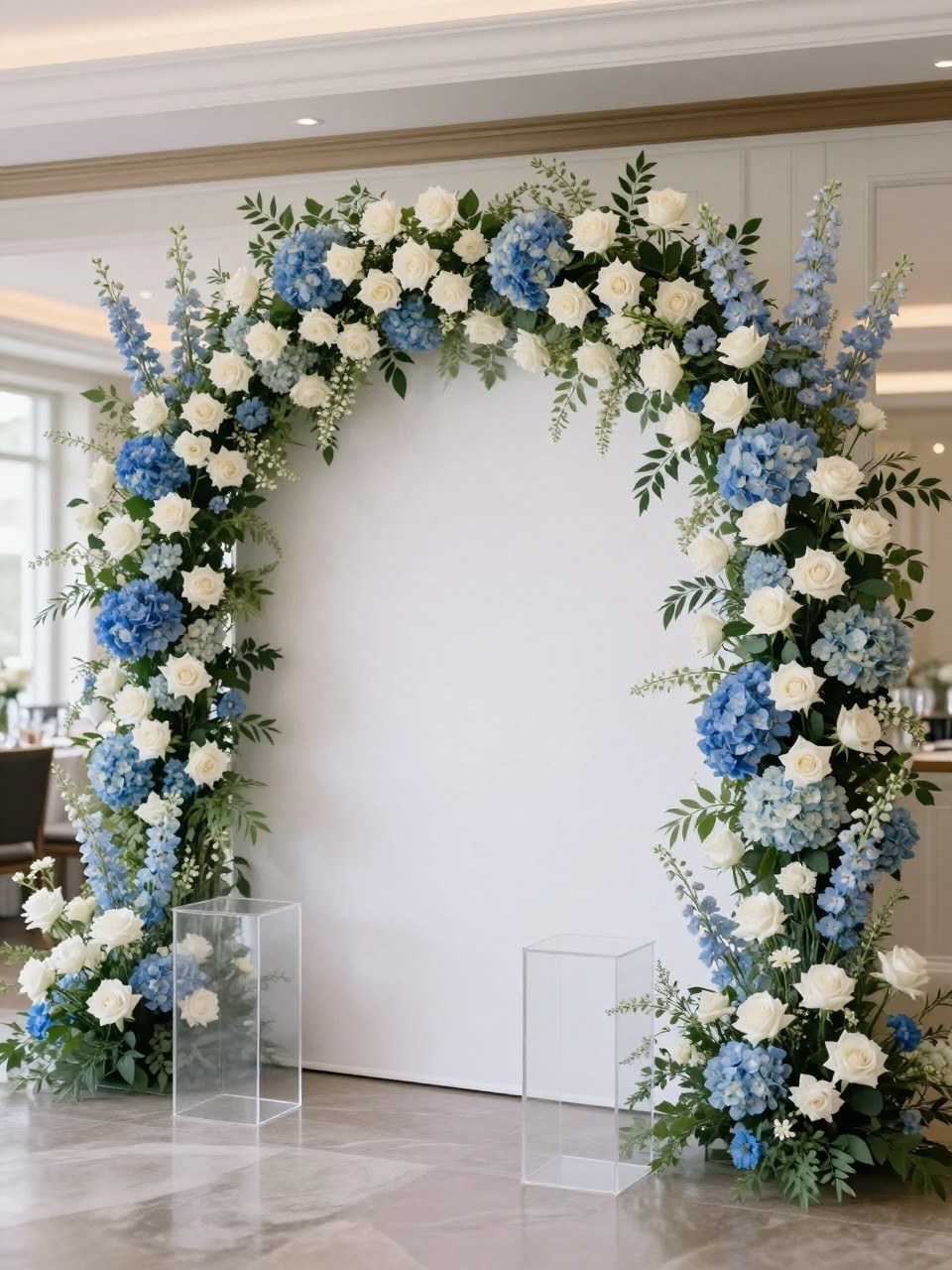 Image Prompt:
A single realistic editorial-style photo of an elegant blue and white flower backdrop with white roses, blue hydrangeas, pale blue delphiniums, soft greenery, and a white circular panel, clear plinths in front, polished indoor party venue, no people, no faces, soft natural lighting, realistic luxury event photo, no text, no watermark.