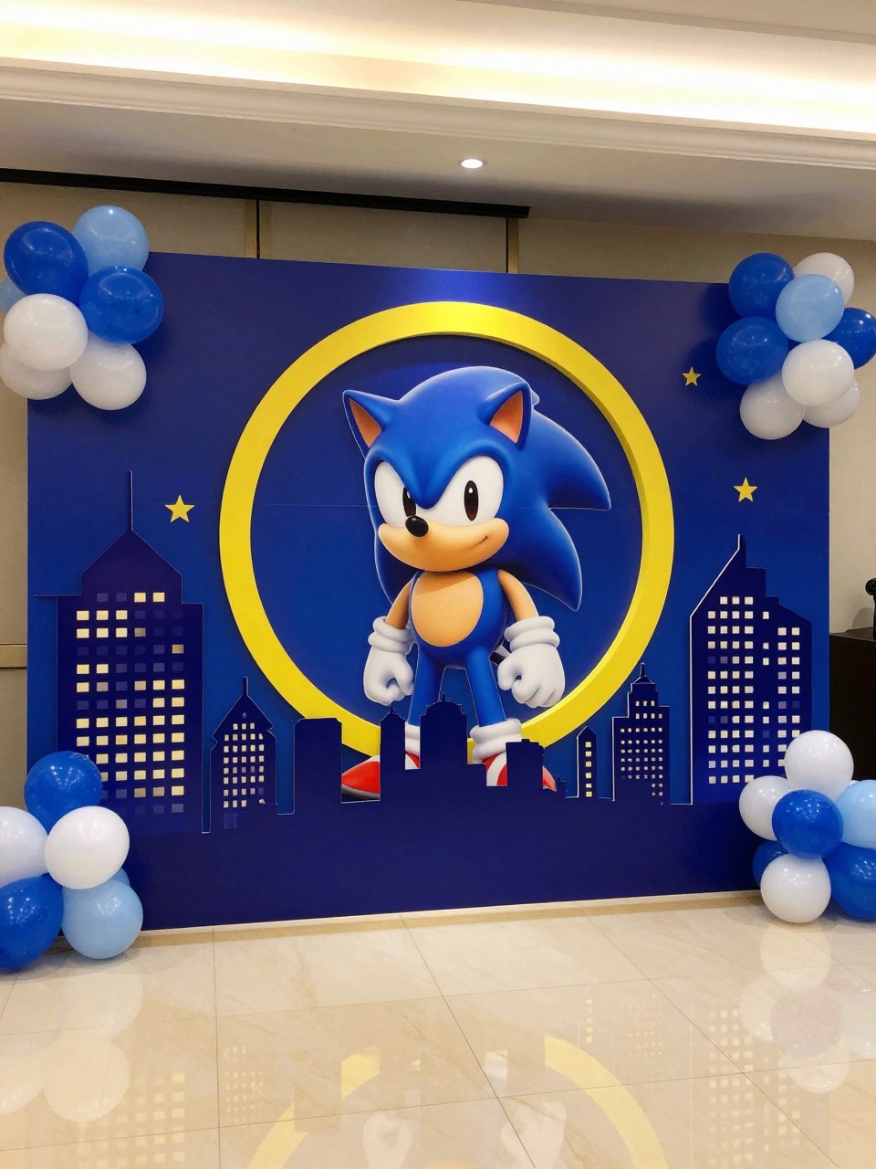 Image Prompt:
A single realistic photo of a creative Sonic-inspired city lights backdrop with deep blue panels, yellow ring accents, small skyline silhouette props, blue and white balloon clusters, red and gold details, glossy indoor floor, evening-style party lighting, no people, no faces, realistic energetic event photography, no readable text, no watermark.