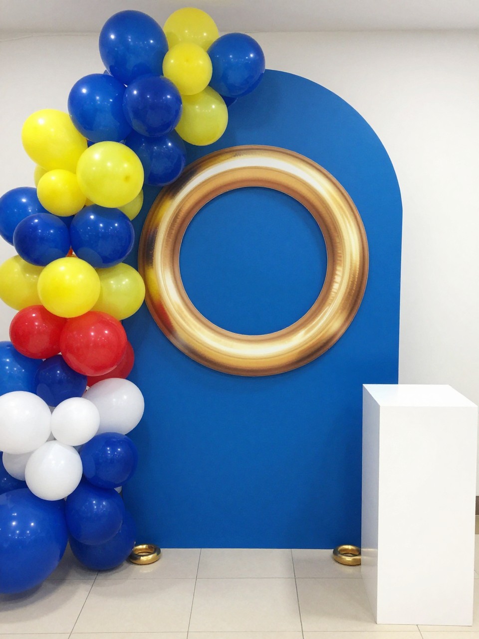 Image Prompt:
A single realistic editorial-style photo of a Sonic-inspired party backdrop with a large gold ring prop as the main focal point, bright blue curved backdrop panel behind it, balloon garland in royal blue, yellow, red, and white flowing around one side, small gold ring props at the base, white plinths, clean indoor birthday party setting, no people, no faces, bright playful lighting, realistic party photography, no text, no watermark.