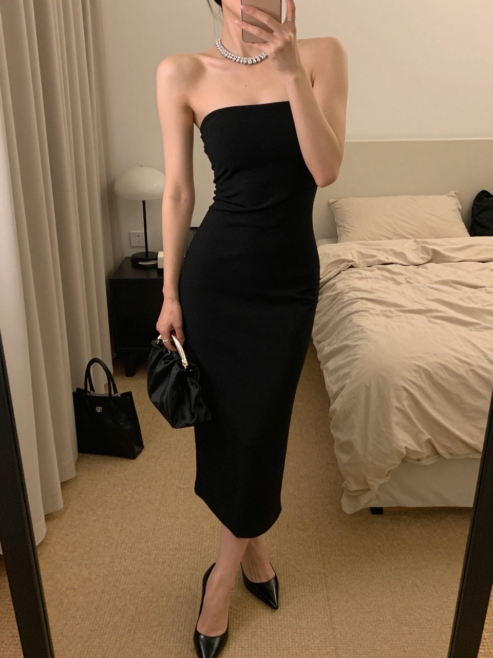 mirror selfie of a woman wearing a black strapless midi dress with sleek heels, a crystal tennis necklace, and a black satin clutch, holding a phone in front of a mirror, cropped at NECK so face doesn't show, with a natural casual selfie pose, in a feminine-themed bedroom, warm moody lighting, including small lived-in details like a tote bag on the floor and soft bedding in the background, latest iPhone photo quality.