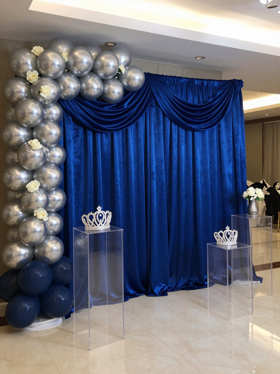 Image Prompt:
A single realistic photo of a royal blue and silver quinceañera backdrop with deep blue velvet draping, silver balloon garland, white roses, crystal-style accents, clear acrylic plinths, silver crown prop, polished indoor event hall, no people, no faces, cool elegant lighting, realistic high-end party photography, no text, no watermark.