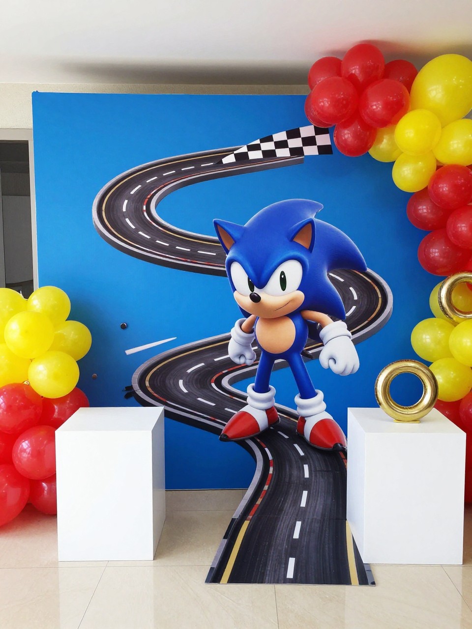Image Prompt:
A single realistic photo of a Sonic-inspired race track party backdrop with a bright blue backdrop panel, black and white checkered racing details, red and yellow balloon clusters, gold ring props, small speed line accents, white cake plinths, indoor birthday party venue, no people, no faces, realistic energetic event photography, no readable text, no watermark.