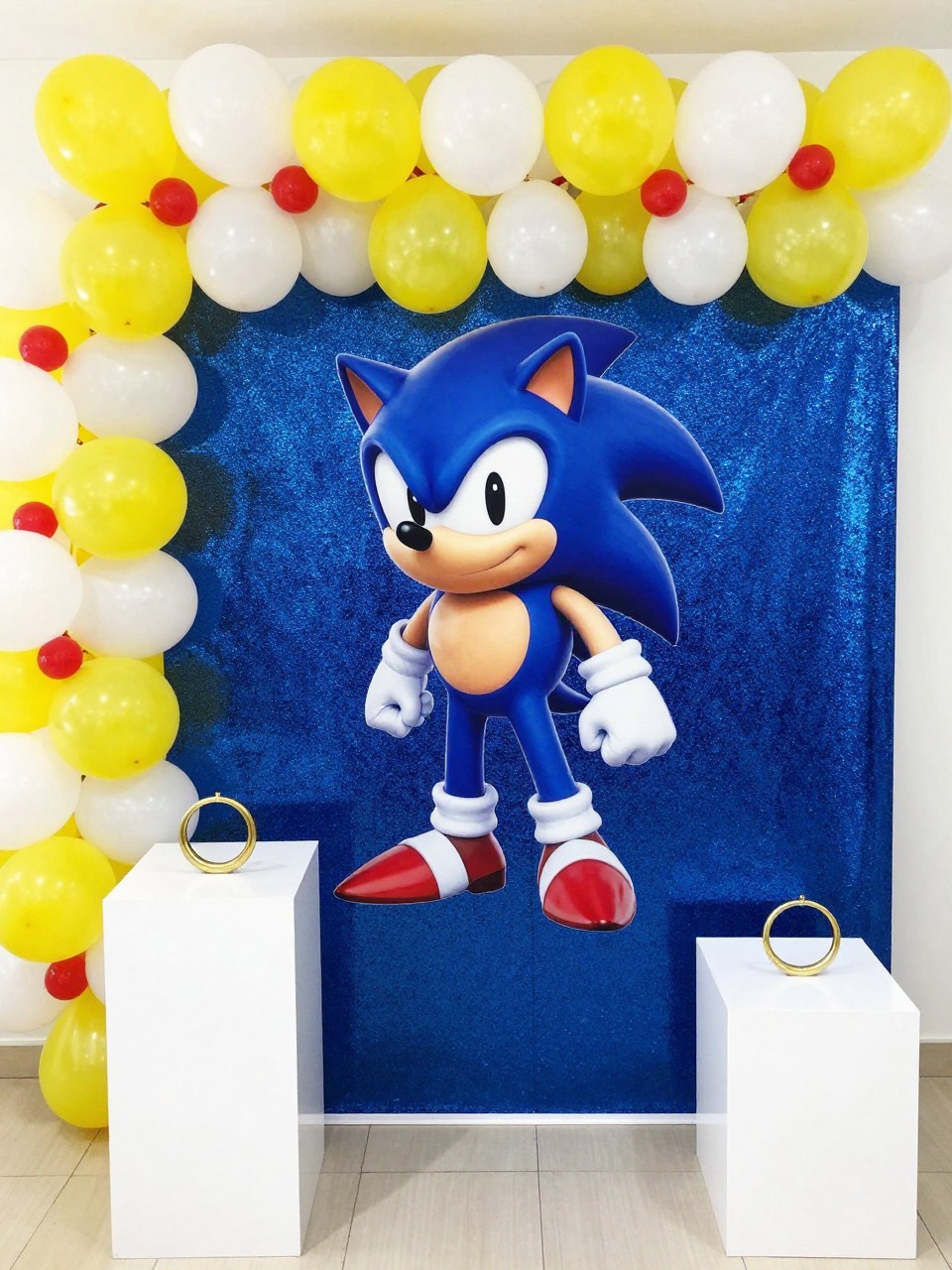 Image Prompt:
A single realistic photo of a Sonic-inspired shimmer wall backdrop with a royal blue shimmer wall, yellow and white balloon garland, red accents, gold ring props at the base, clean white pedestal stands, bright indoor birthday party setting, no people, no faces, realistic glossy event photography, no text, no watermark.