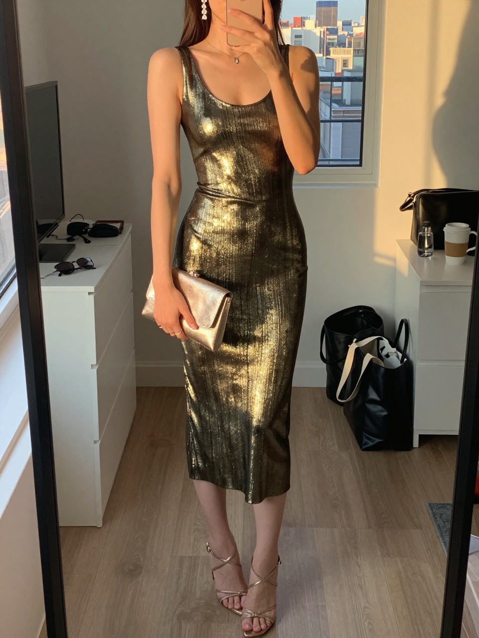 mirror selfie of a woman wearing a metallic gold midi dress with strappy stiletto heels, a champagne metallic clutch, and crystal drop earrings, holding a phone in front of a mirror, cropped at NECK so face doesn't show, with a natural casual selfie pose, in a city apartment, evening golden hour, including small lived-in details like sunglasses on a dresser, a tote bag on the floor, or a coffee mug nearby, latest iPhone photo quality.