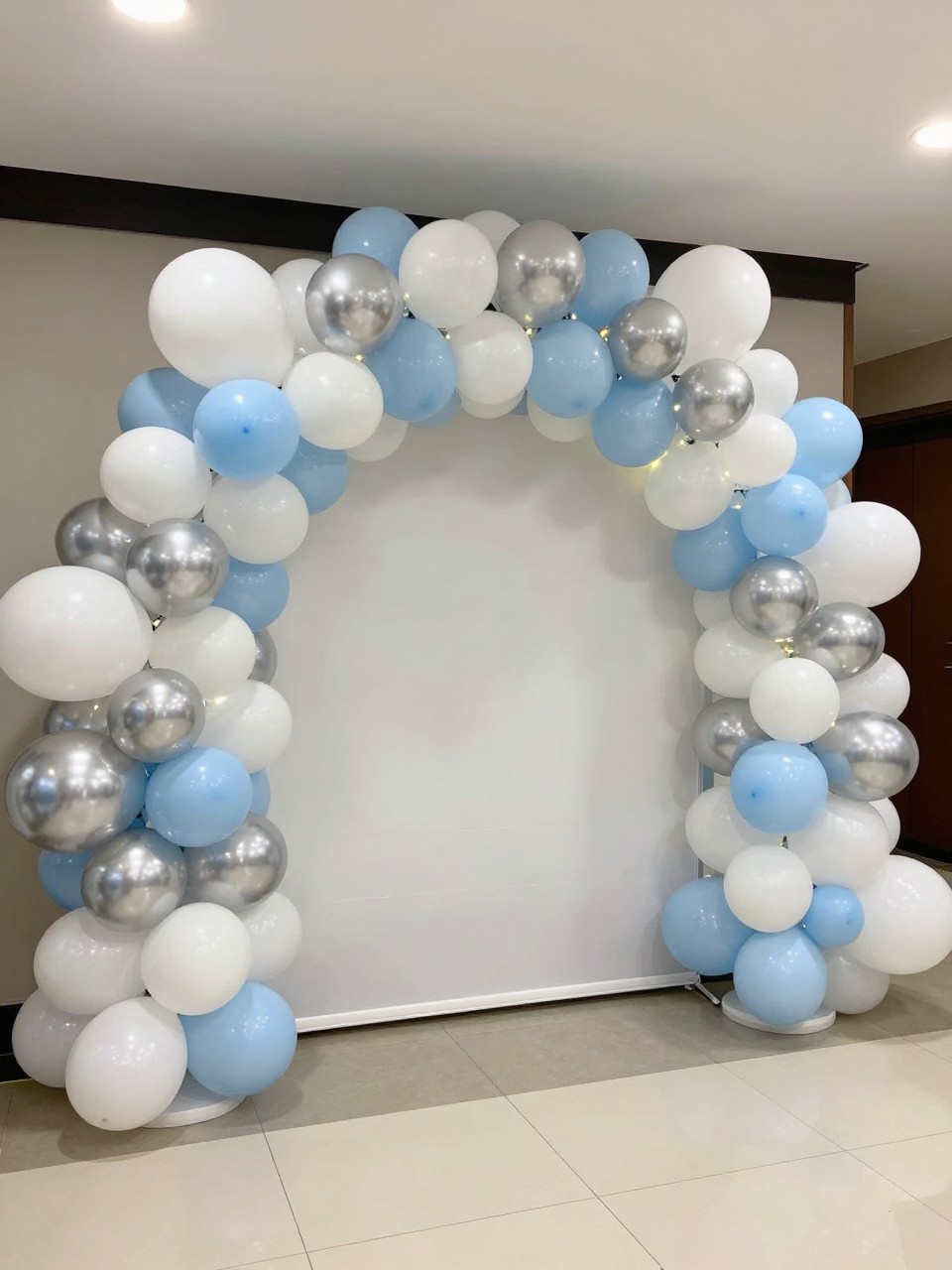 Image Prompt:
A single realistic photo of a balloon tunnel backdrop at a party entrance, with layered balloons in white, soft blue, silver, and pearl tones forming a rounded tunnel shape, small lights tucked inside the balloon clusters, clean event floor, modern indoor celebration space, no people, no faces, realistic event photography, no text, no watermark.