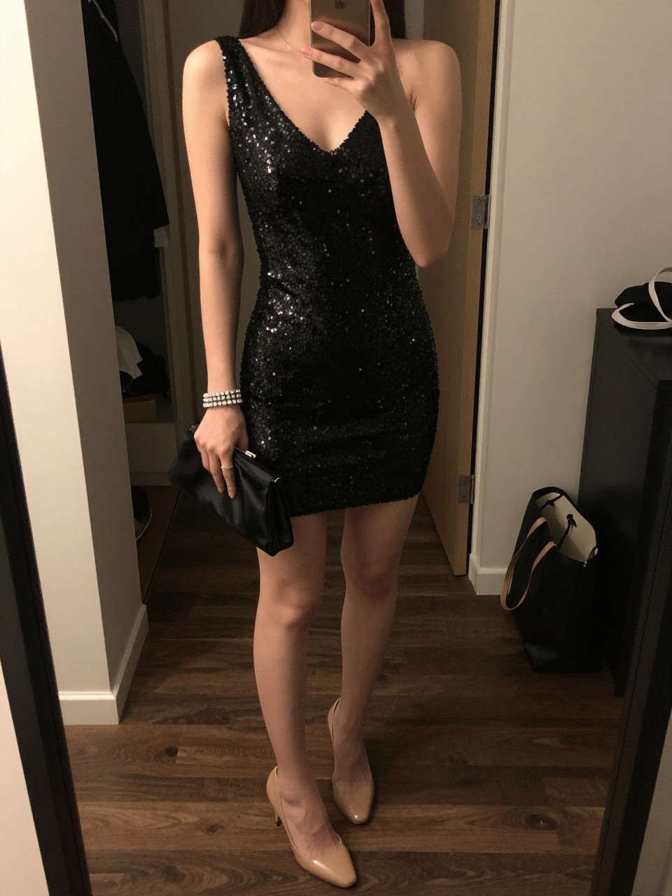 mirror selfie of a woman wearing a black sequin mini dress with nude barely-there heels, a black satin clutch, and a crystal tennis bracelet, holding a phone in front of a mirror, cropped at NECK so face doesn't show, with a natural casual selfie pose, in a city apartment, warm moody lighting, including small lived-in details like an open closet behind and a tote bag on the floor, latest iPhone photo quality.