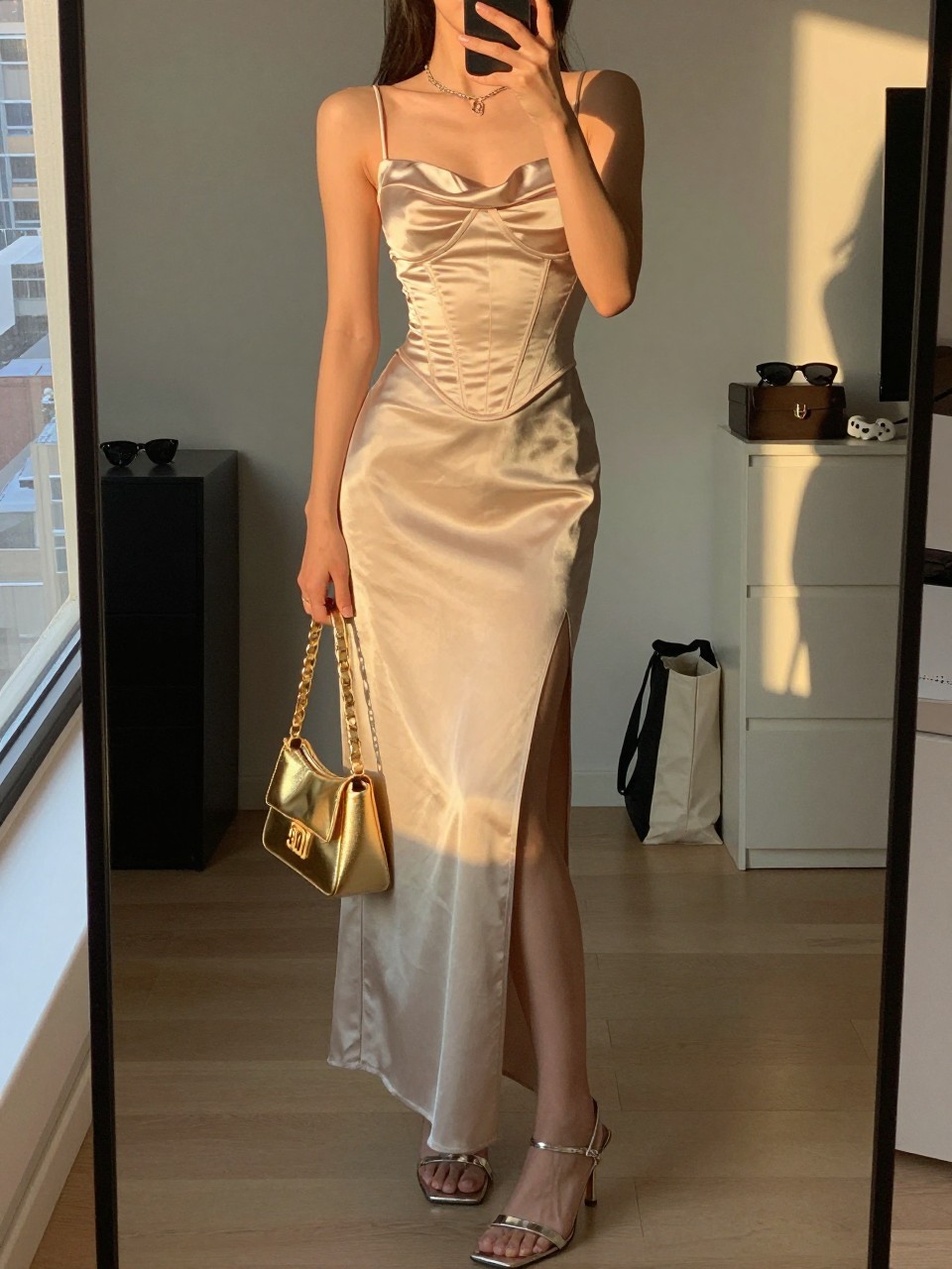 mirror selfie of a woman wearing a satin corset top with a matching high-slit maxi skirt, metallic heeled sandals, and a metallic gold mini bag, holding a phone in front of a mirror, cropped at NECK so face doesn't show, with a natural casual selfie pose, in a city apartment, evening golden hour, including small lived-in details like sunglasses on a dresser and a tote bag on the floor, latest iPhone photo quality.