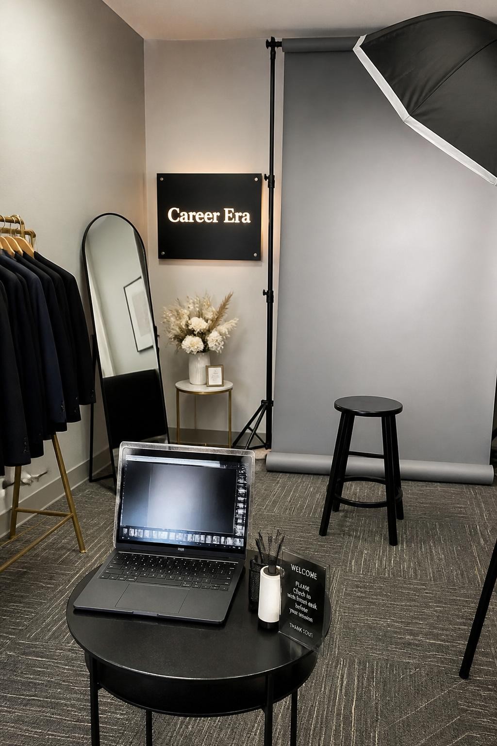 A realistic DSLR photo of a modern graduation headshot lounge with a clean backdrop, stool, softbox-style lighting, laptop on a side table, blazer rack, lint roller, mirror, neutral flowers, and a small sign that says “Career Era,” polished indoor setup, realistic photography station, no people.