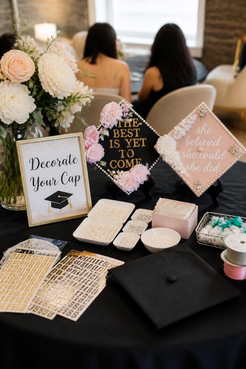 A realistic DSLR photo of a classy graduation cap decorating station with faux flowers, pearl stickers, letter stickers, ribbons, glue dots, small trays, sample decorated caps, and a sign that says “Decorate Your Cap,” two guests sitting in the background from behind and softly out of focus, bright indoor party lighting, realistic creative event setup.