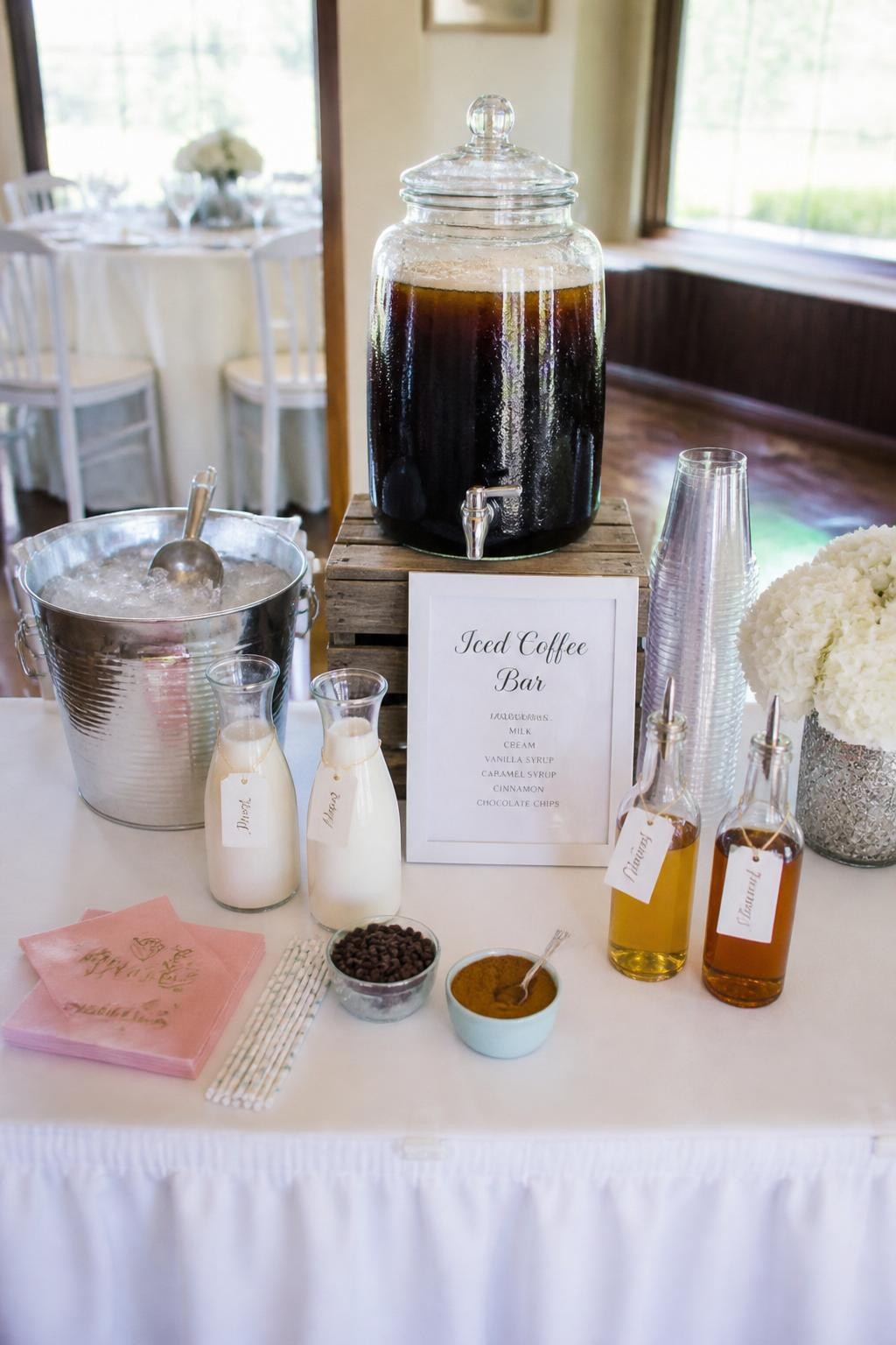 A realistic DSLR photo of a bright bridal shower iced coffee bar, clear glass drink dispenser filled with cold brew, ice bucket with silver scoop, clear cups stacked neatly, glass bottles of milk and cream, vanilla and caramel syrup bottles, small bowls of chocolate chips and cinnamon, white flowers, pale pink napkins, sunny party room, realistic styling, no people.