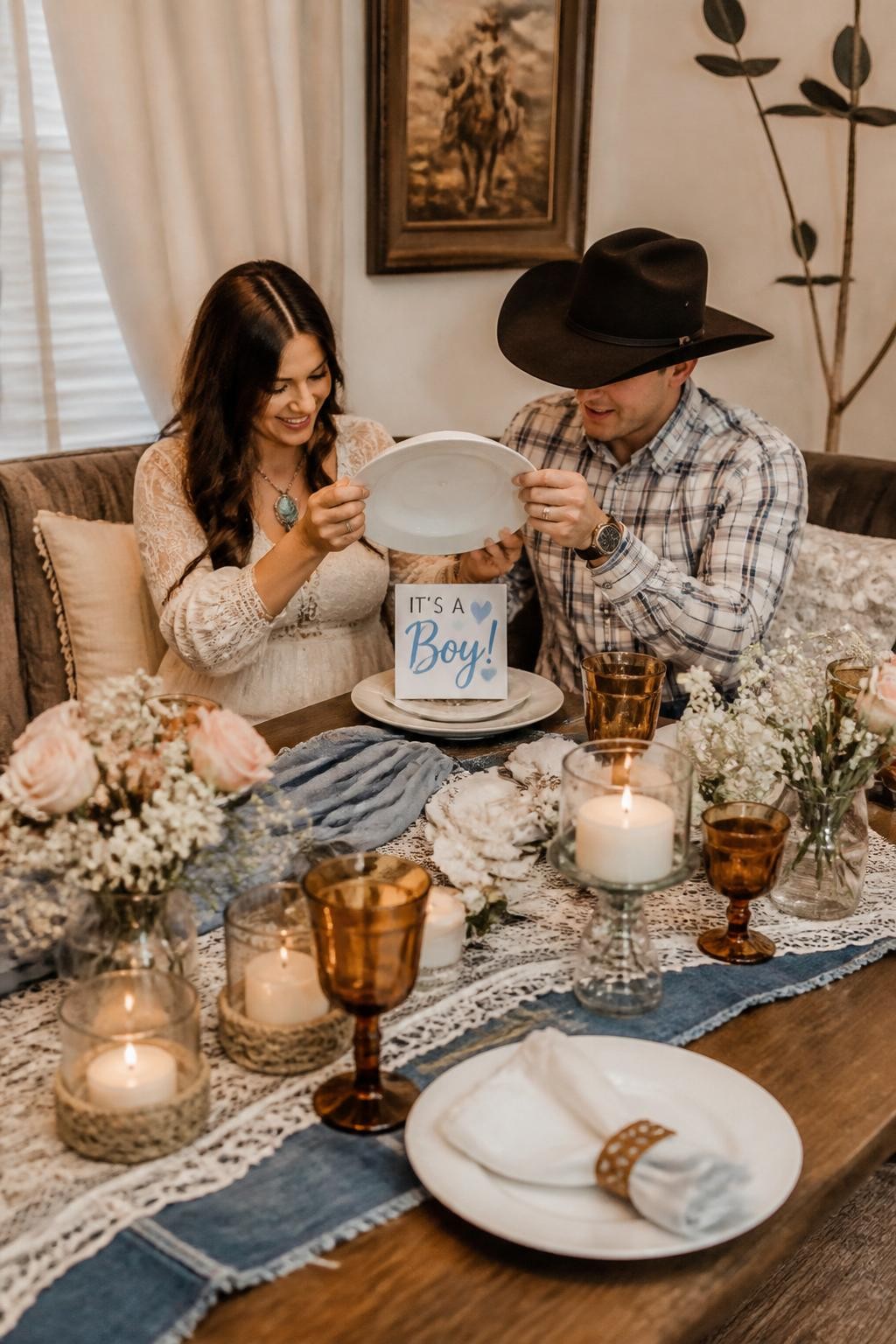 A realistic editorial photo of an elegant western gender reveal dinner table, layered denim and lace runner, wooden table, white plates, amber glasses, soft florals, candles, parents lifting a covered plate or place card with the reveal message, refined western home decor, warm indoor lighting, vertical DSLR photography