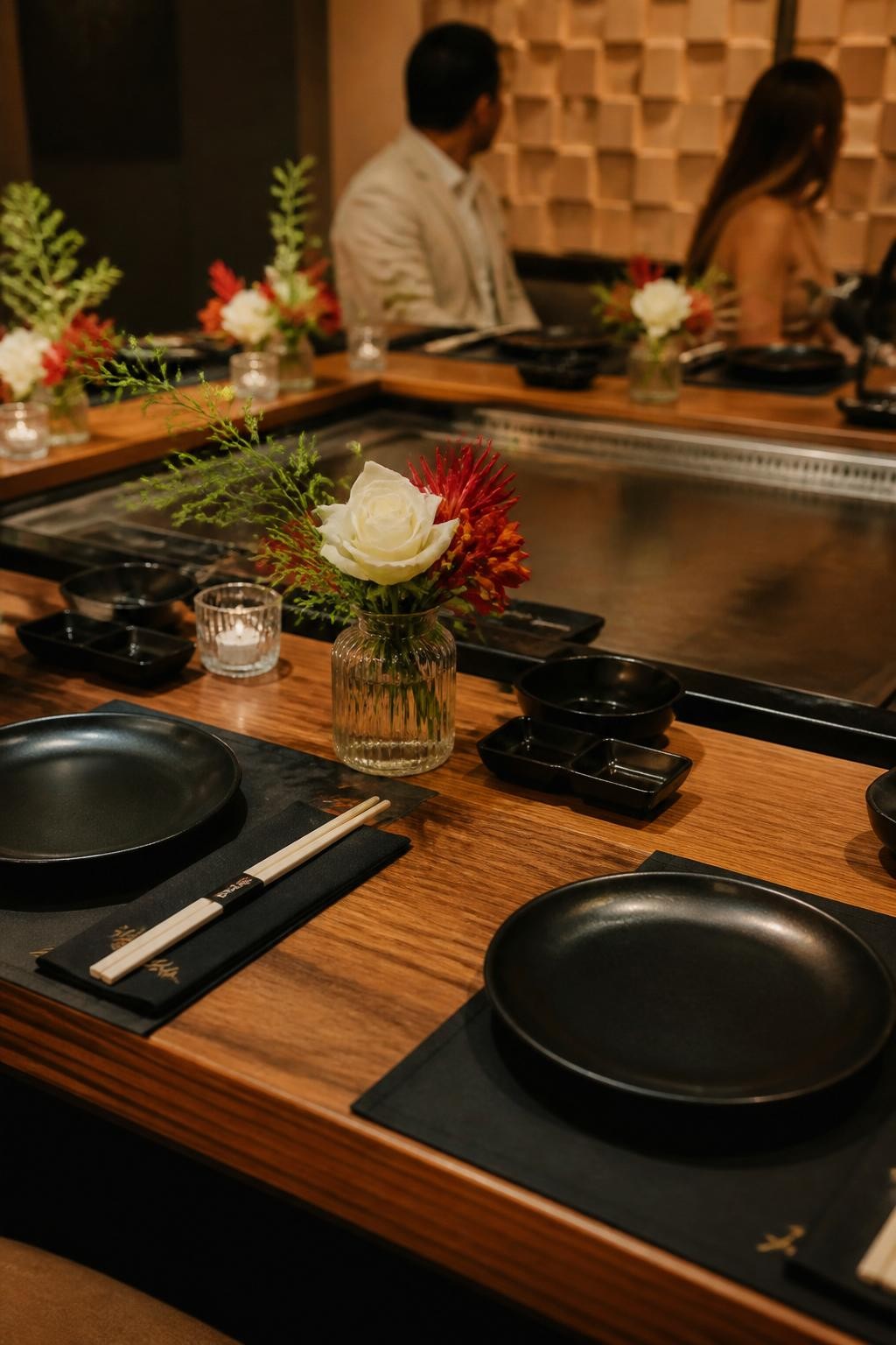 Image Prompt:**
A realistic DSLR photo of a stylish hibachi party table arranged around a central grill area, with black plates, chopsticks, small sauce bowls, low floral accents in red and white, fresh greenery, clean wood textures, modern warm lighting, two guests slightly out of focus seated at the table and turned away from the camera, editorial event styling.**