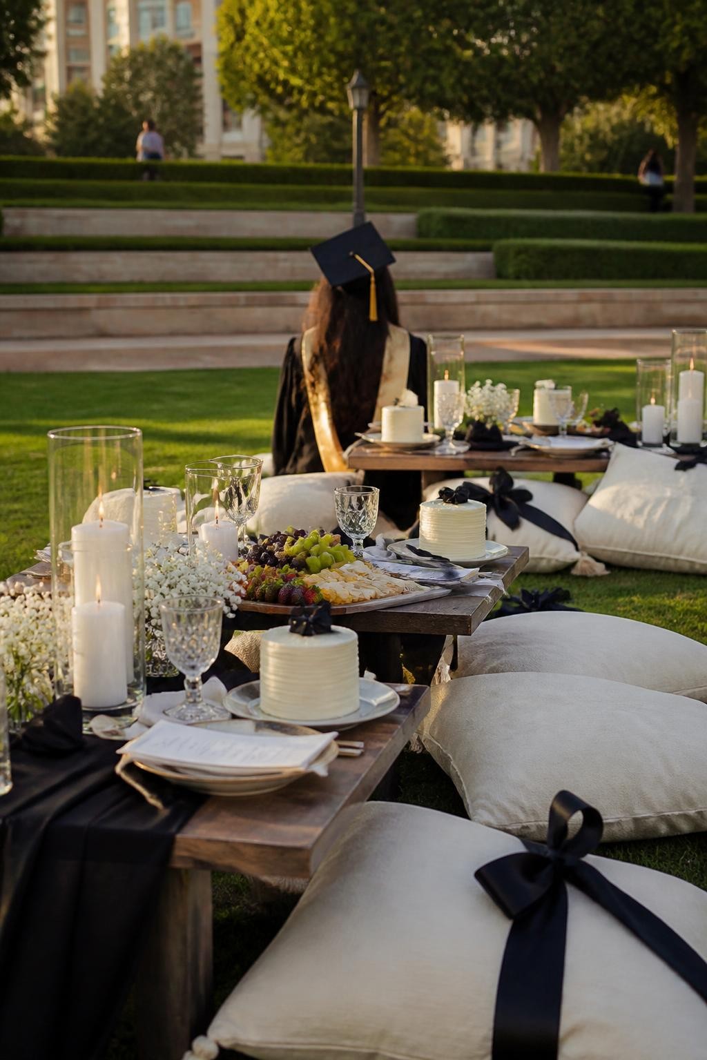 A realistic DSLR photo of an outdoor college graduation picnic with low tables, ivory cushions, black satin bows, glassware, mini cakes, fruit boards, candles in hurricanes, and a graduate in a black gown sitting with their back turned in the soft-focus background, golden hour lawn setting, elegant modern picnic styling.