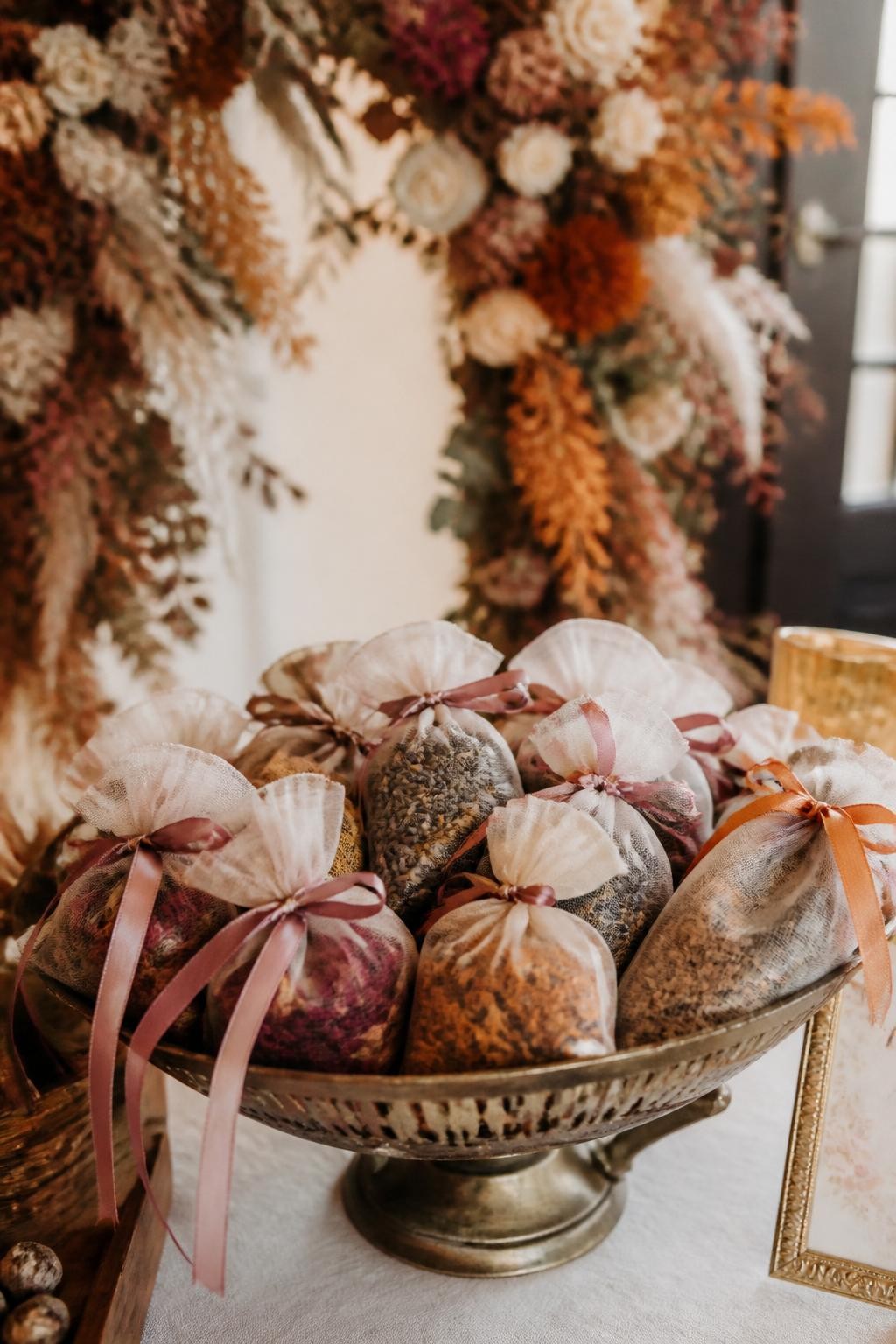 Realistic DSLR photo of small sheer organza and linen sachets filled with dried lavender, rose petals, marigold, and cedar chips, tied with mauve and rust ribbons, displayed in a vintage brass bowl on a wedding favor table, dried floral arch softly blurred in the background, romantic autumn indoor lighting.