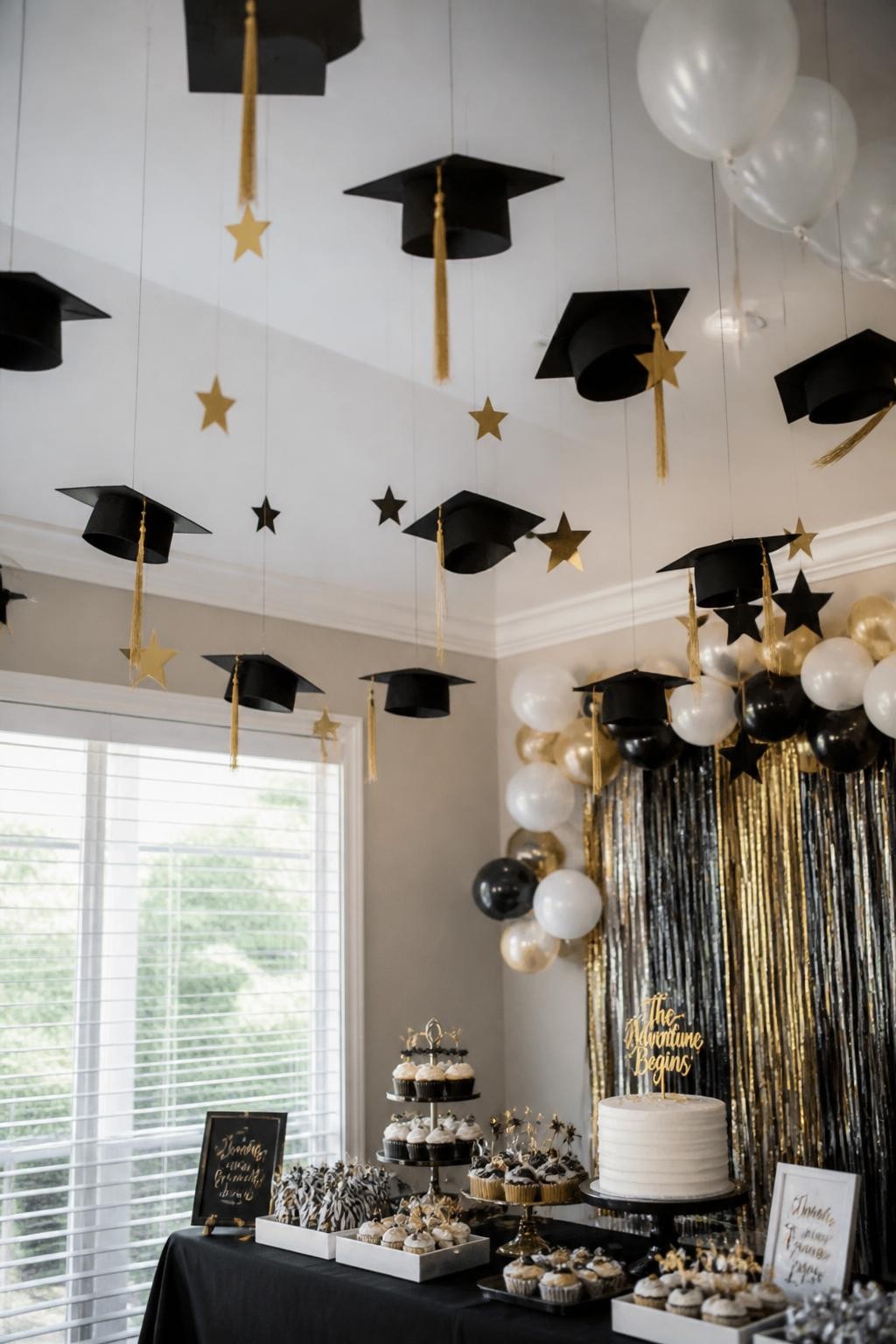 A realistic DSLR photo of an indoor graduation party ceiling installation with paper graduation caps, hanging tassels, metallic stars, clear fishing line, soft white balloons, and a dessert table underneath with cupcakes and a small cake, modern black, white, and gold palette, bright natural light, realistic event decor, no people.