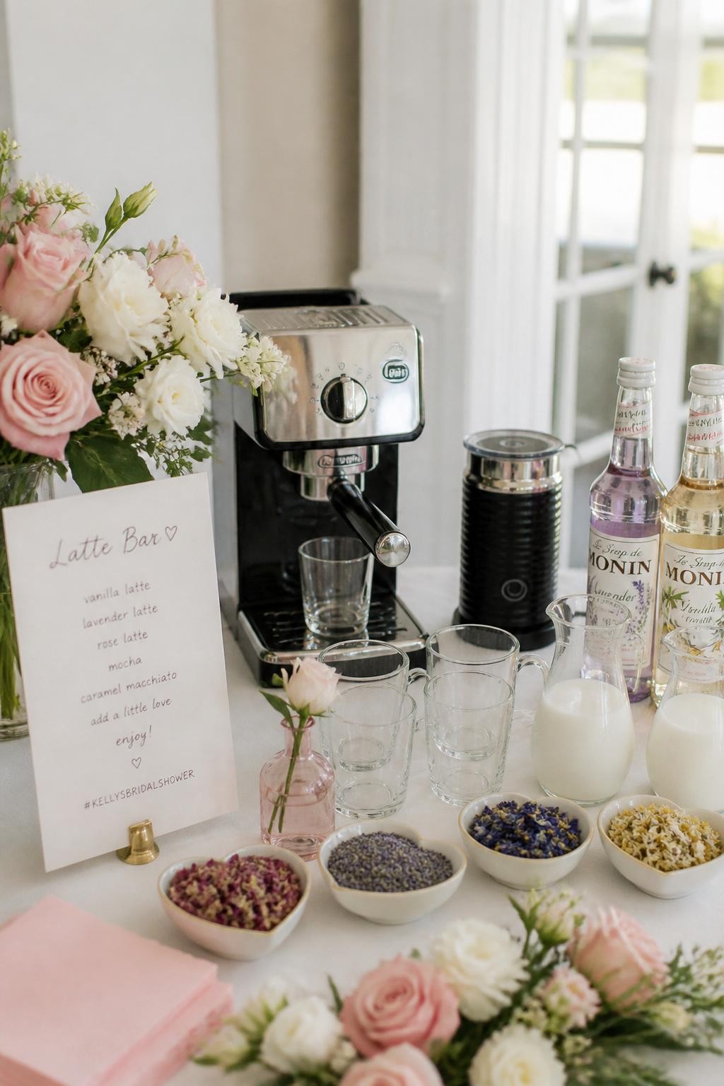 A realistic DSLR photo of a bridal shower latte bar with a compact espresso machine, milk frother, clear glasses, small pitchers of milk, dried edible flower garnish in tiny bowls, lavender syrup bottle, vanilla syrup bottle, white and pink flowers around the table, handwritten latte menu card, bright airy room, soft daylight, realistic party styling, no people.