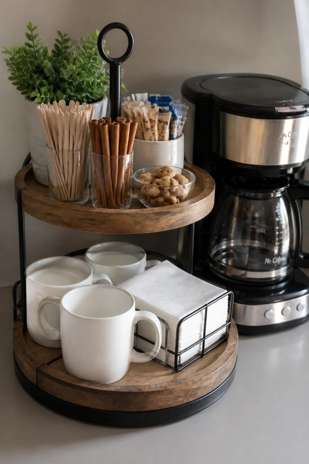 A realistic DSLR photo of a small coffee bar using a two-tier stand, lower tier holding mugs and napkins, upper tier holding sugar packets, stirrers, cinnamon sticks, and mini cookies, coffee carafe beside it, neutral kitchen counter, small faux greenery, soft morning light, organized budget-friendly coffee station, no people.