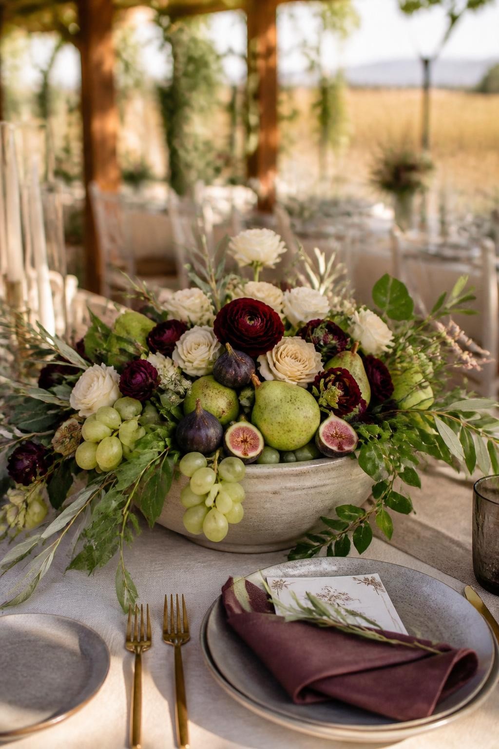 Realistic DSLR photo of a large shallow ceramic bowl centerpiece filled with green pears, fresh figs, grapes, burgundy ranunculus, cream roses, olive branches, and trailing greenery, placed on a fall wedding table with stoneware plates, gold flatware, linen runner, and warm vineyard reception background, elegant natural light.