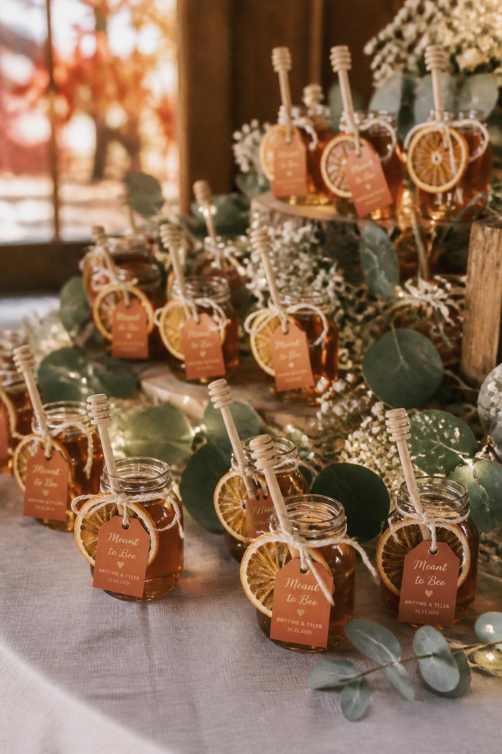 Realistic DSLR photo of tiny glass honey jars with golden honey, wooden dipper sticks, dried orange slices tied to the neck with cream cotton string, and small burnt-orange tags, displayed on a linen-covered wedding favor station with eucalyptus, baby’s breath, and warm fairy lights, soft fall wedding background.