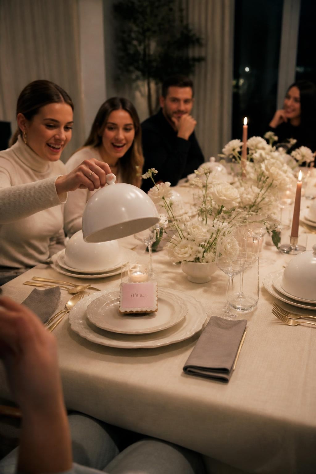 A realistic photo of an elegant home dining table set for a gender reveal dinner, cream tablecloth, sculptural plates, clear glassware, folded napkins, soft florals, guests seated around the table, covered mini cloches on each plate, one cloche lifted to reveal a dessert or card in one soft reveal color, cozy evening lighting, editorial dinner party photography