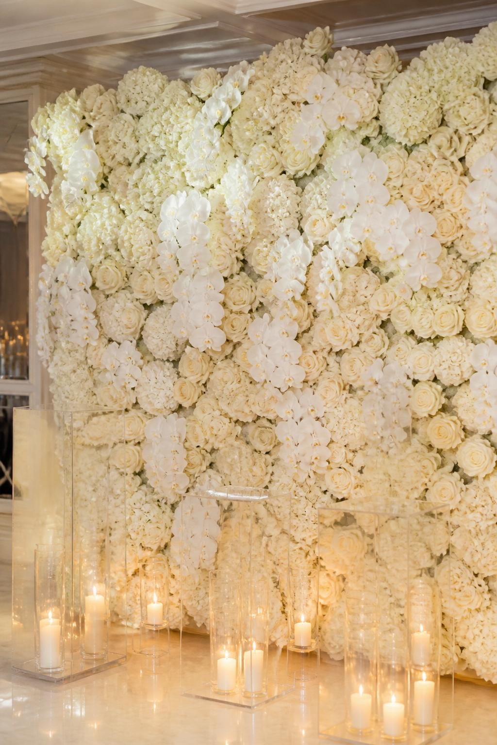 Image Prompt:
A single realistic editorial-style photo of a gorgeous flower backdrop with a full wall of white roses, ivory hydrangeas, and soft white orchids, clear acrylic plinths in front, warm glass candle holders at the base, polished indoor event venue, no people, no faces, soft diffused lighting, realistic luxury event photography, no text, no watermark.